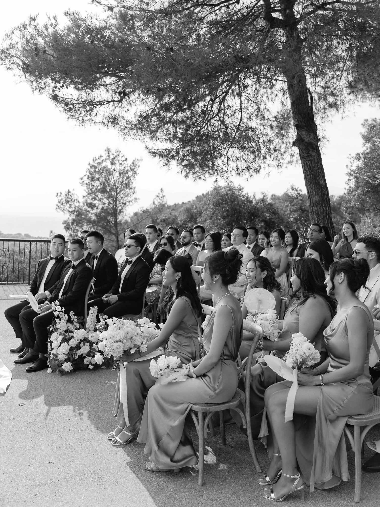 Black and white outdoor wedding ceremony on terrace with bridal party and guests under pine tree