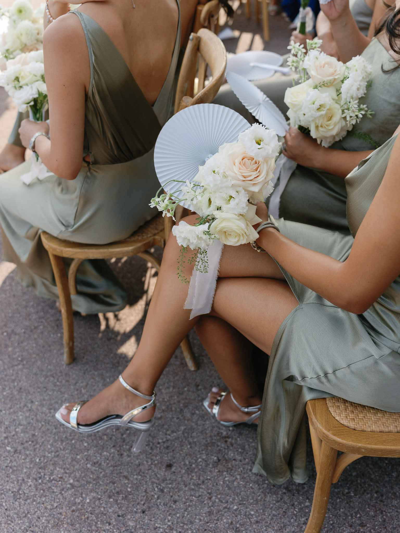 Bridal party with sage green dresses holding white and peach bouquets seated on wooden chairs indoors