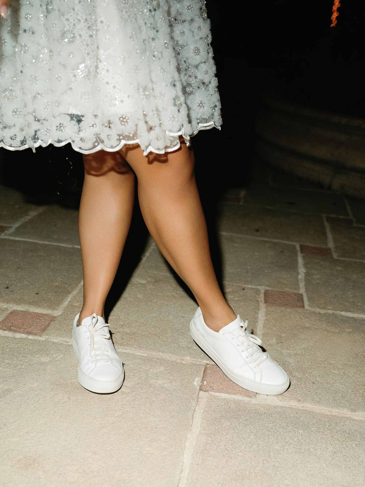 Bride in white lace dress and canvas sneakers on stone pavement at evening wedding reception