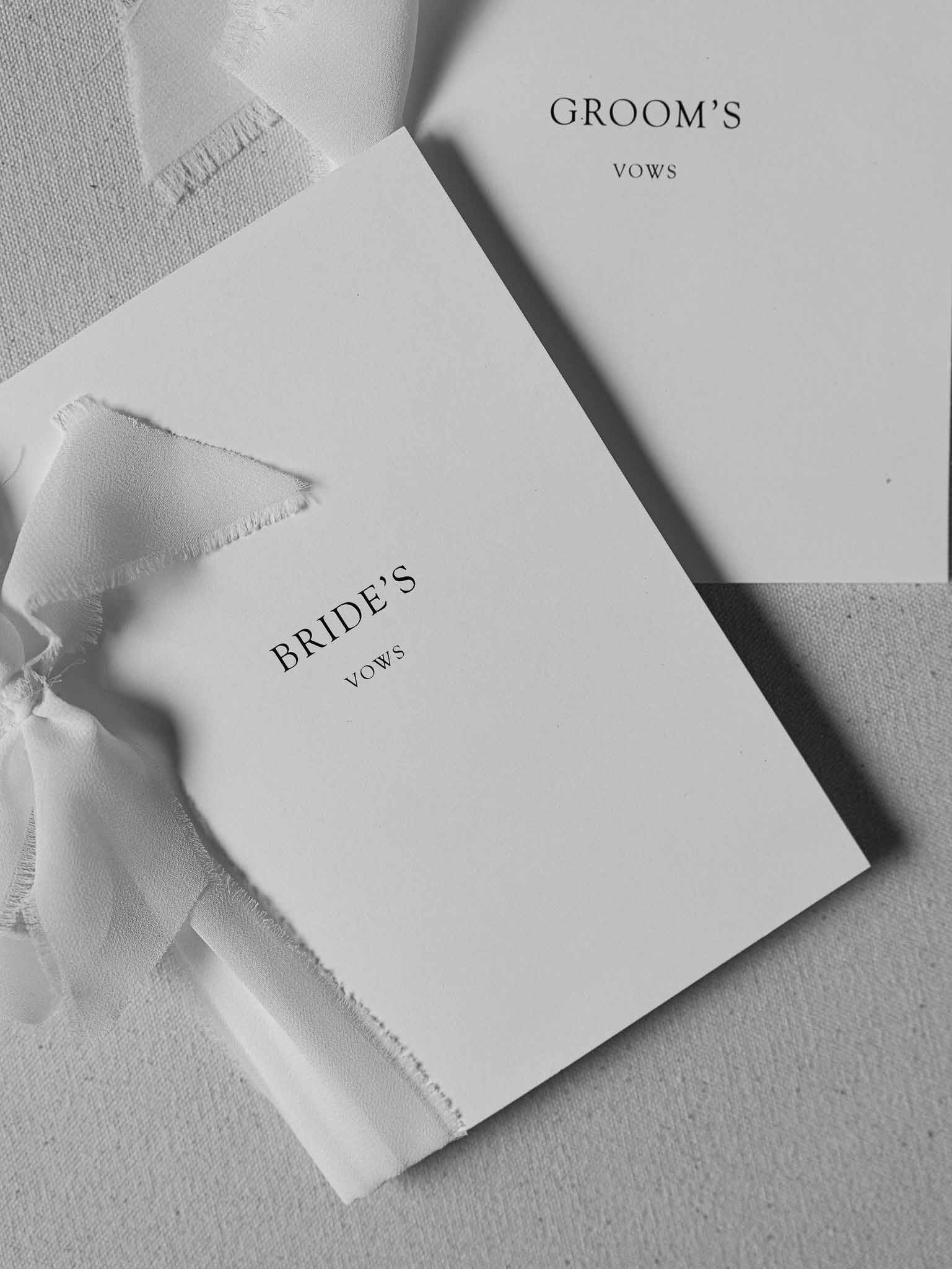 Black and white flat lay of bride and groom vow booklets with silk ribbon bows on cream linen