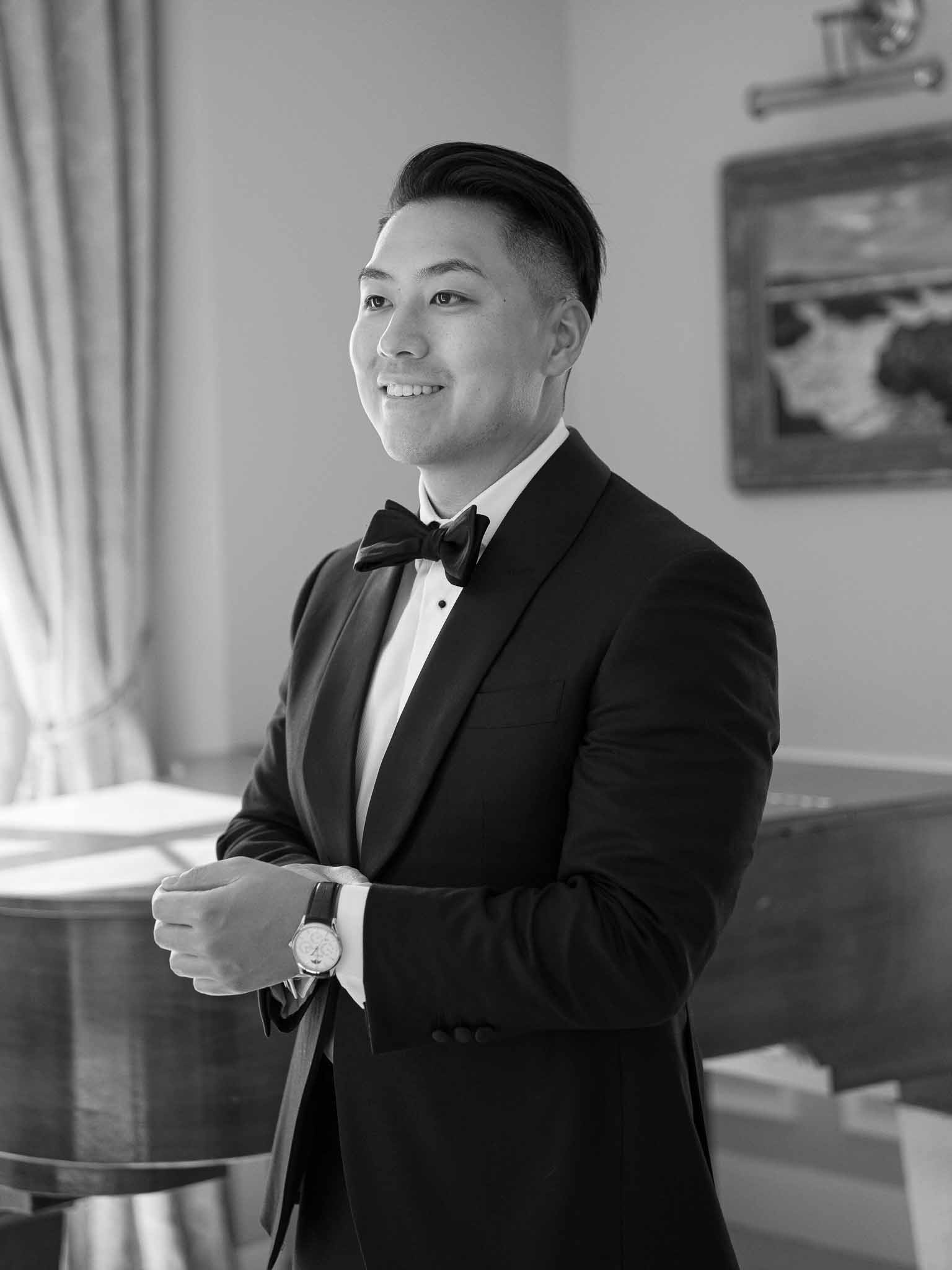 Groom in black tuxedo and bow tie during getting ready preparations in bright interior room