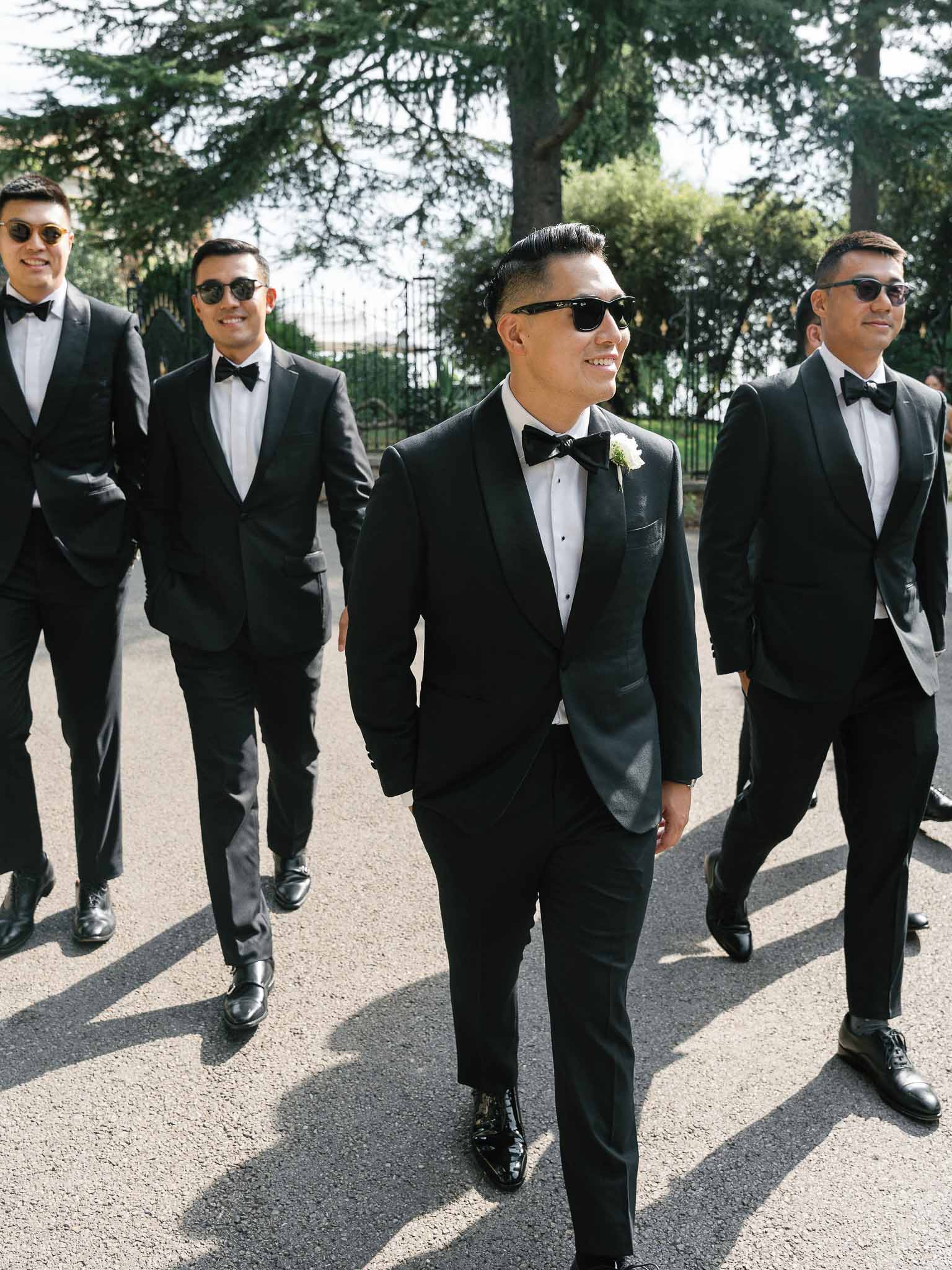Groom and groomsmen in black tuxedos walking along garden path at outdoor wedding venue