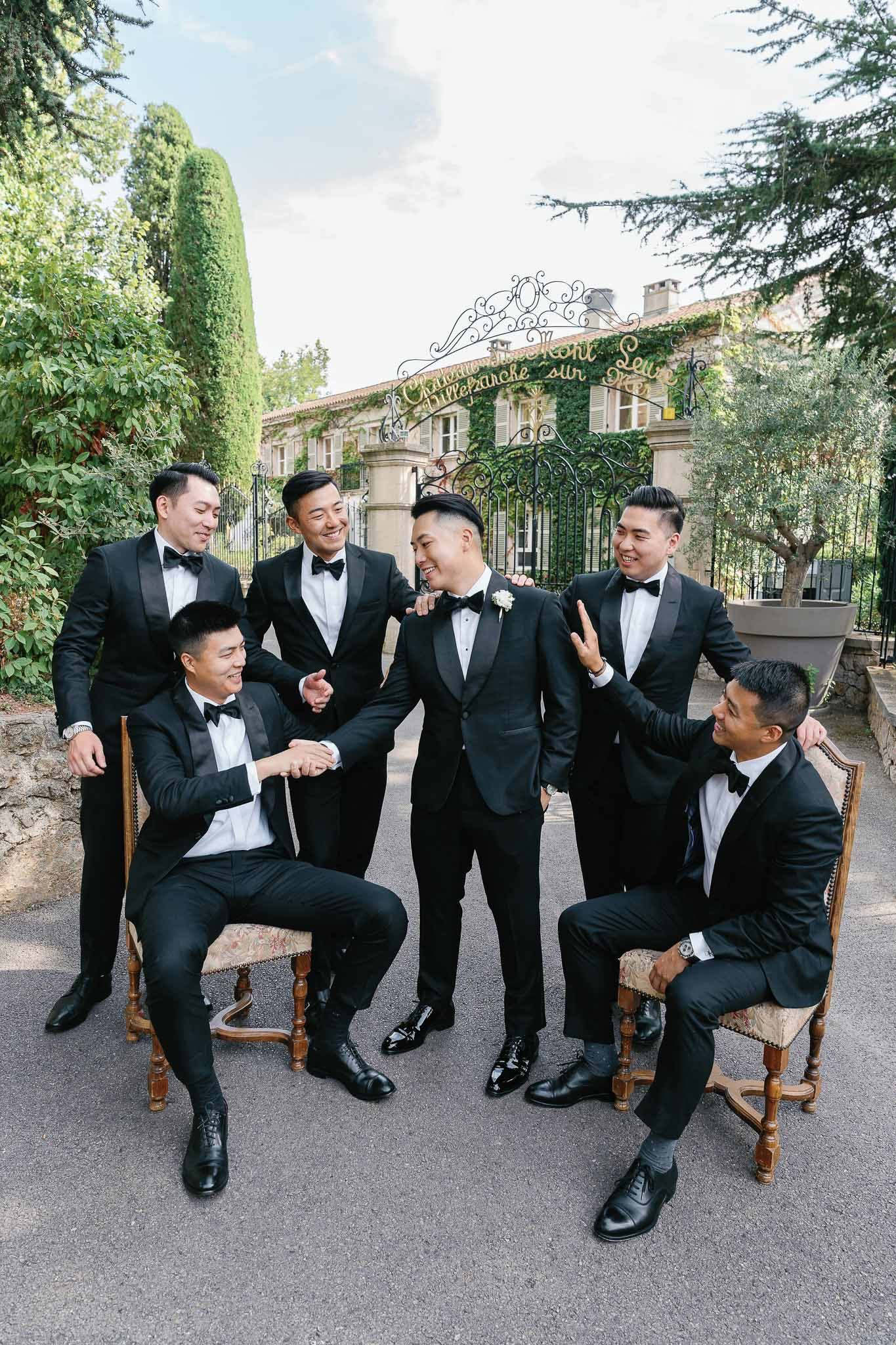 Groom and groomsmen in black tuxedos posing for group portrait in ivy-covered mansion courtyard