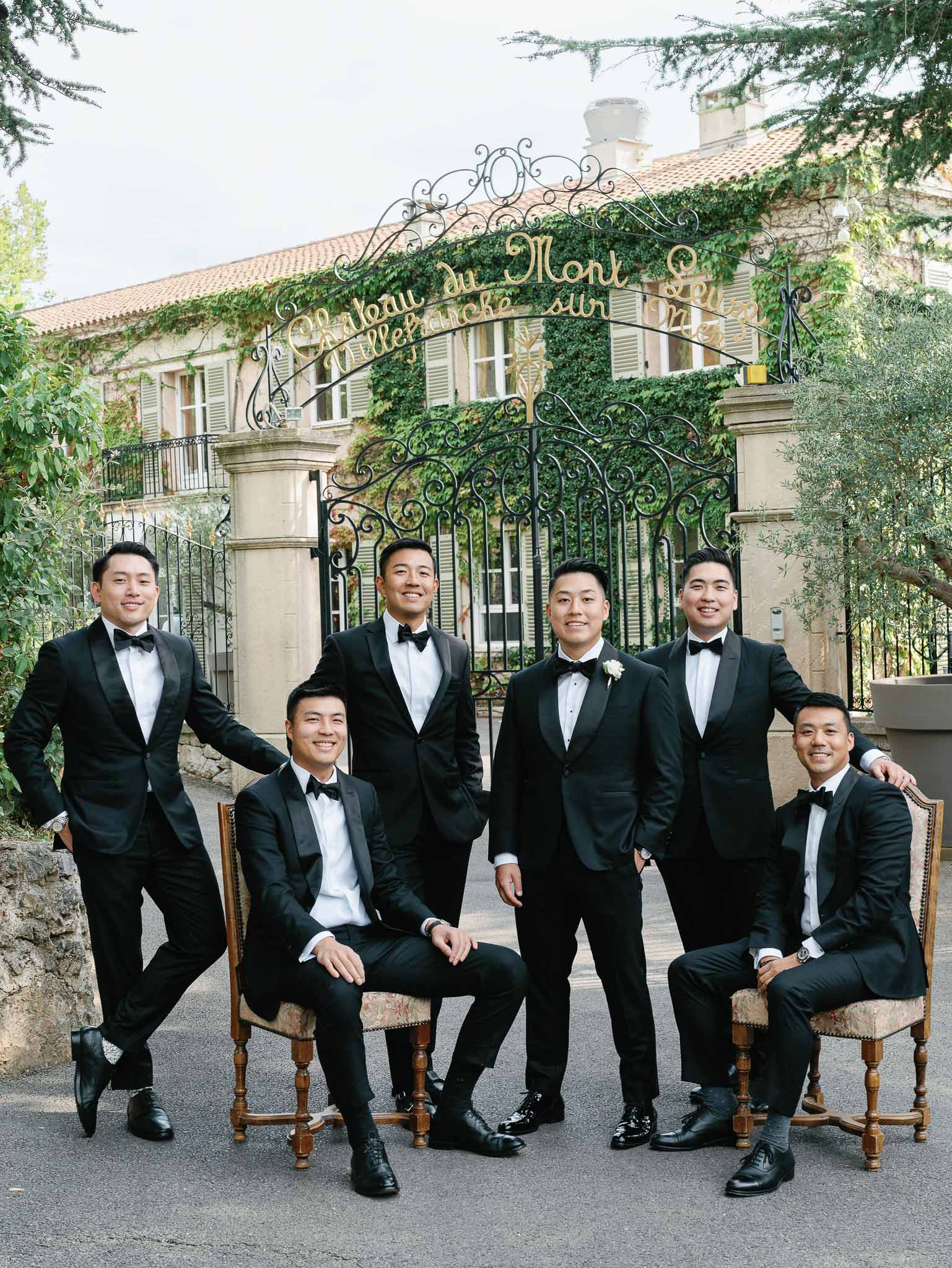 Groom and groomsmen in black tuxedos posing for formal portrait at French estate with wrought-iron gate