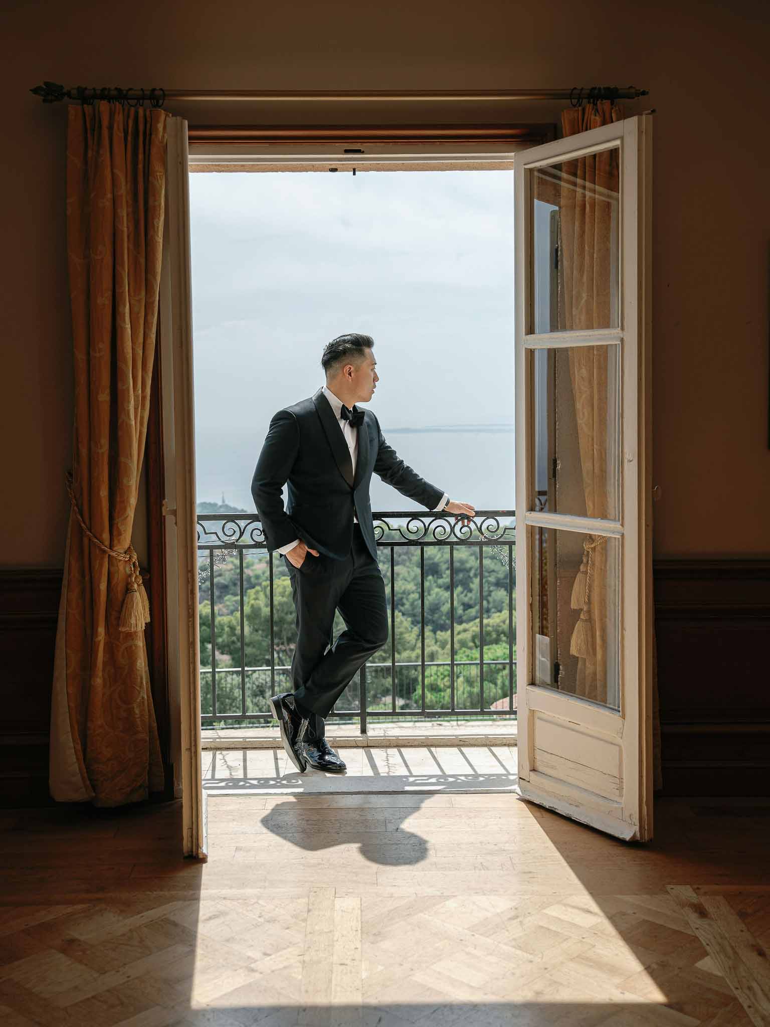 Groom in black tuxedo on balcony overlooking water view at chateau venue