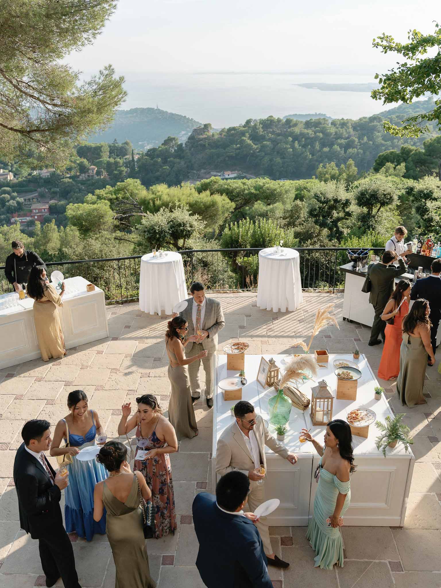 Cocktail hour on stone terrace overlooking Mediterranean landscape with guests at white high-top tables