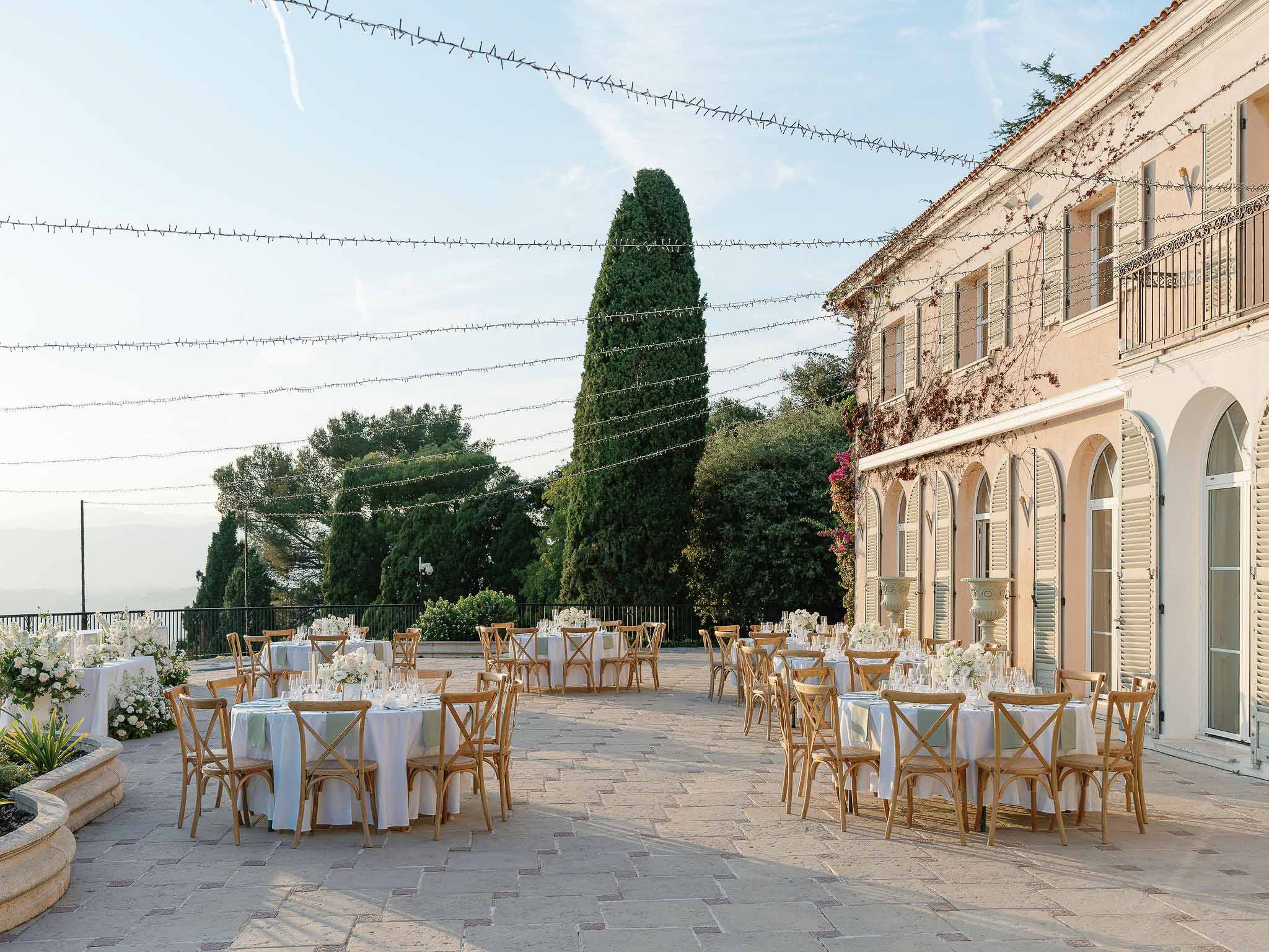 Reception terrace setup with round tables at classical French chateau with string lights and formal gardens