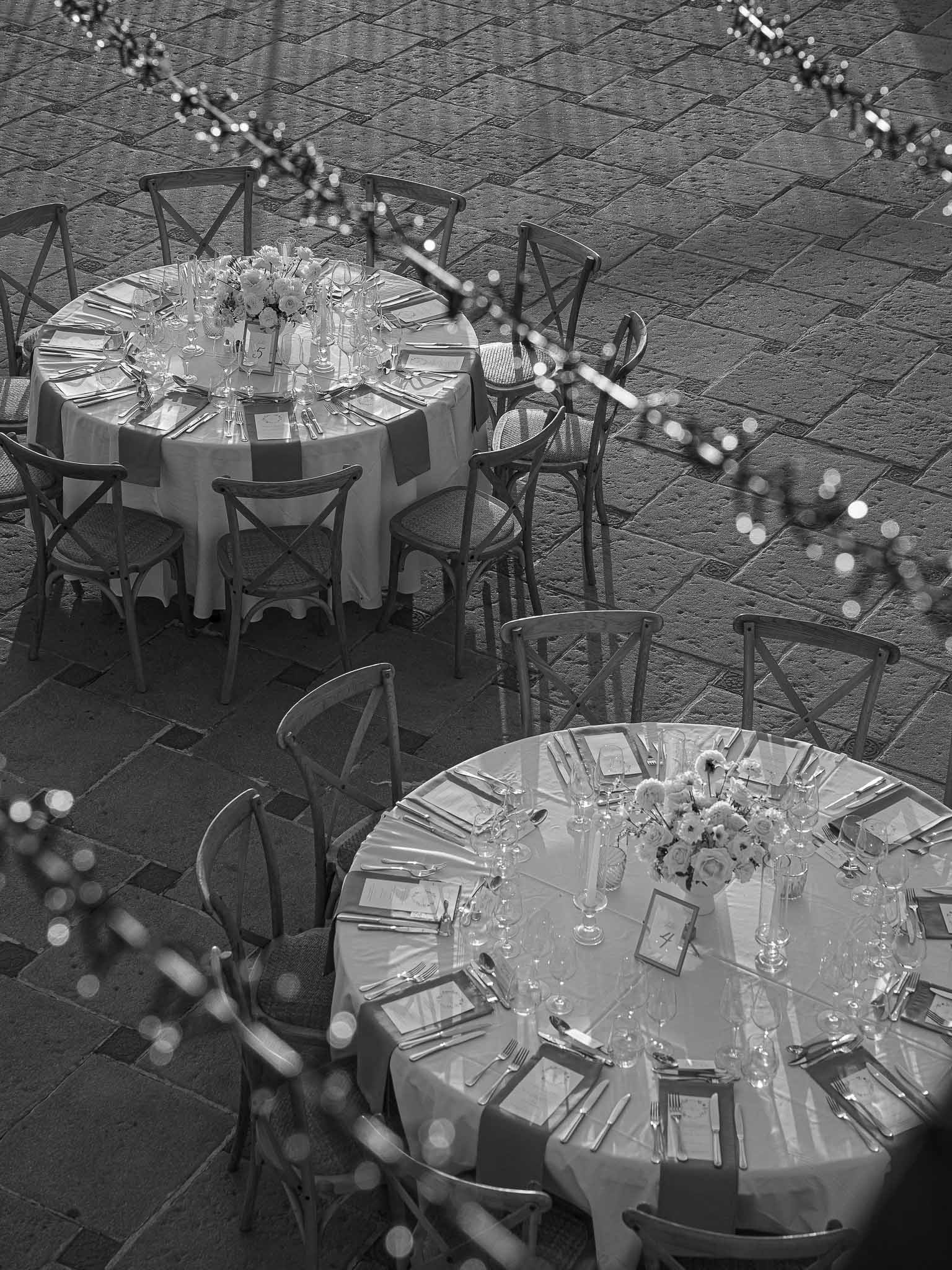 Black and white overhead view of elegant round table settings in outdoor courtyard with herringbone paving