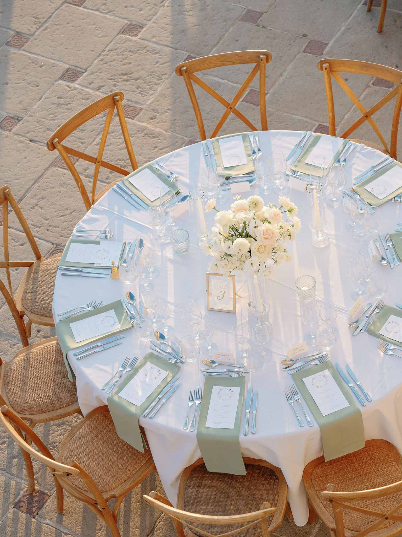 Overhead round table with sage napkins, blush rose centerpiece, gold table number, and cross-back chairs