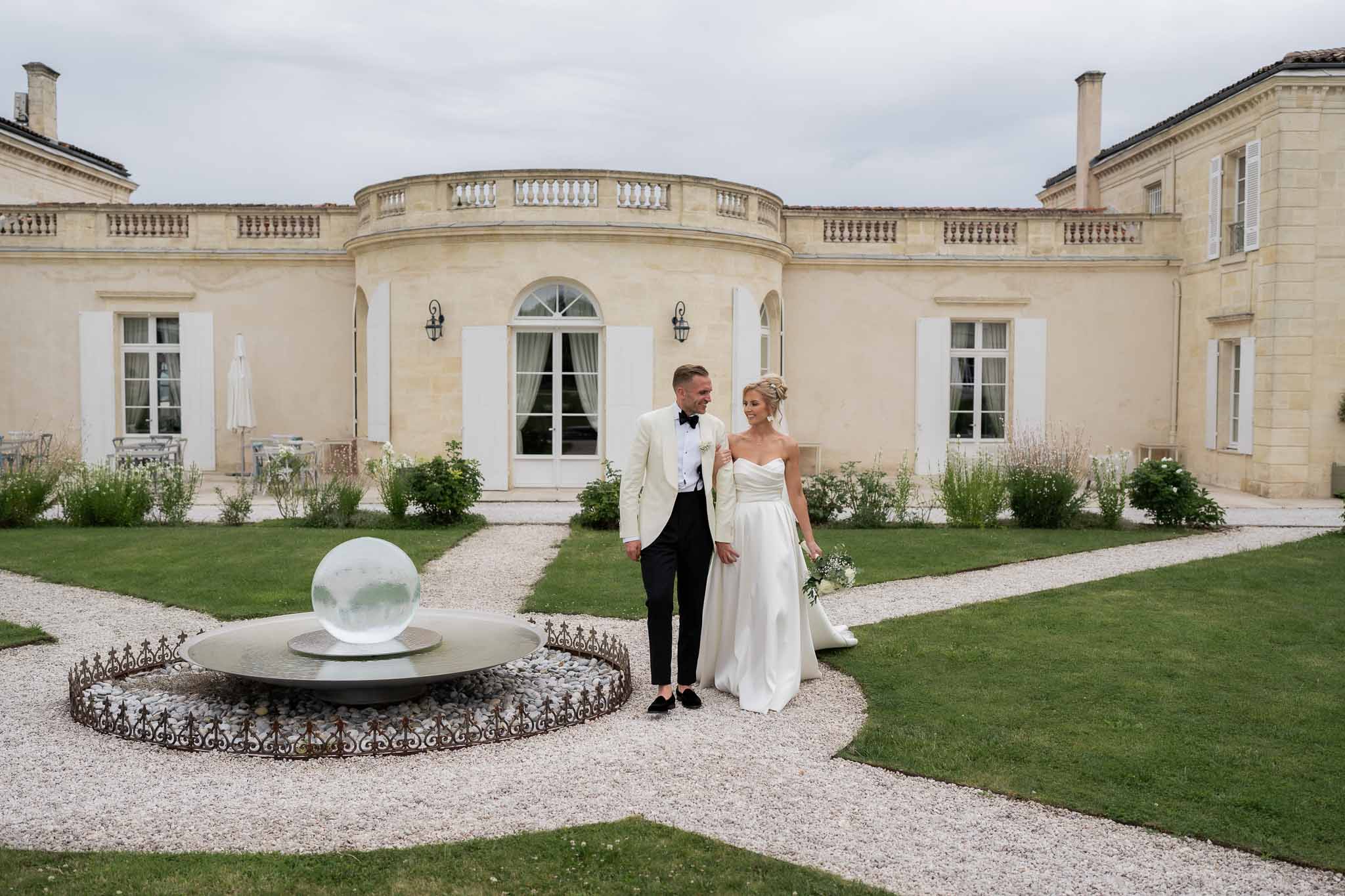 Bride and groom pose together in courtyard of classical stone château with water feature