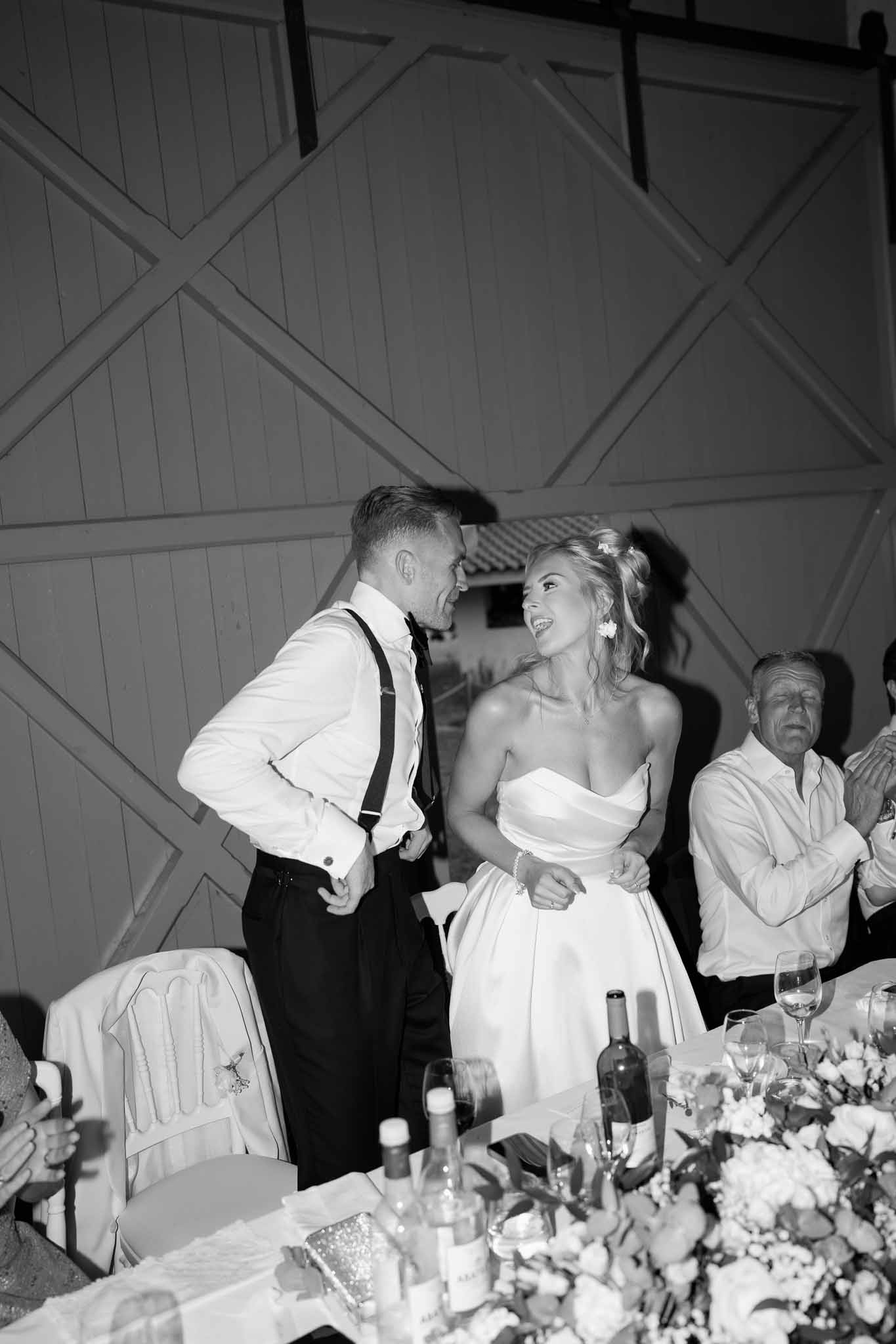 Bride and groom interacting joyfully at reception dinner table with guests looking on