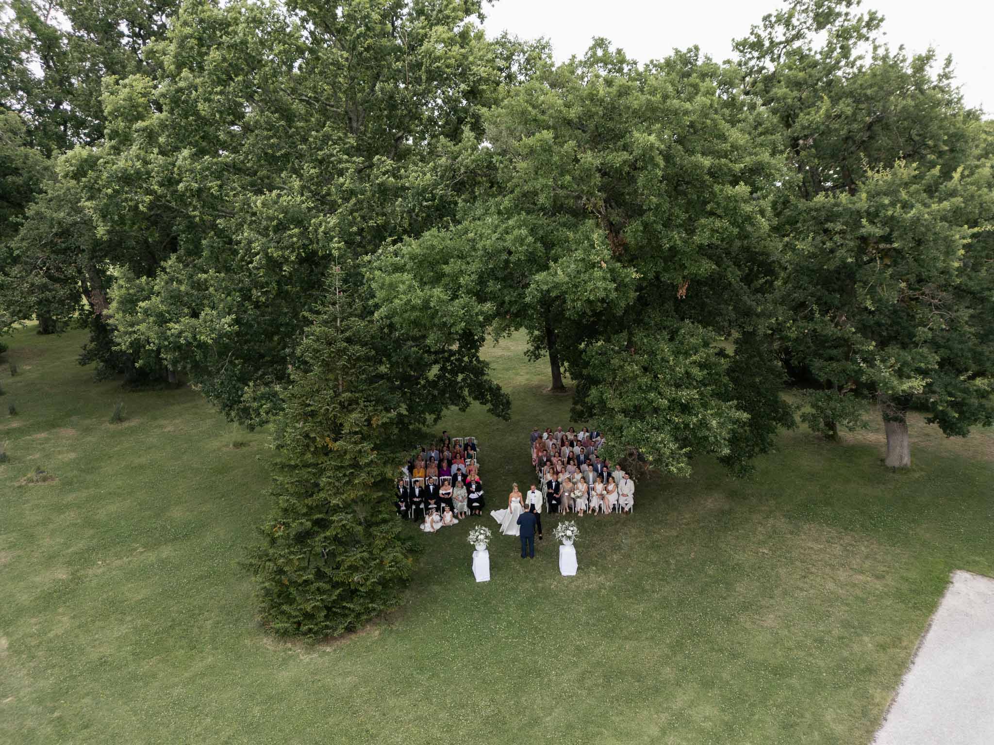 Aerial drone view of outdoor wedding ceremony on lawn with 80-100 guests in white chairs under mature trees