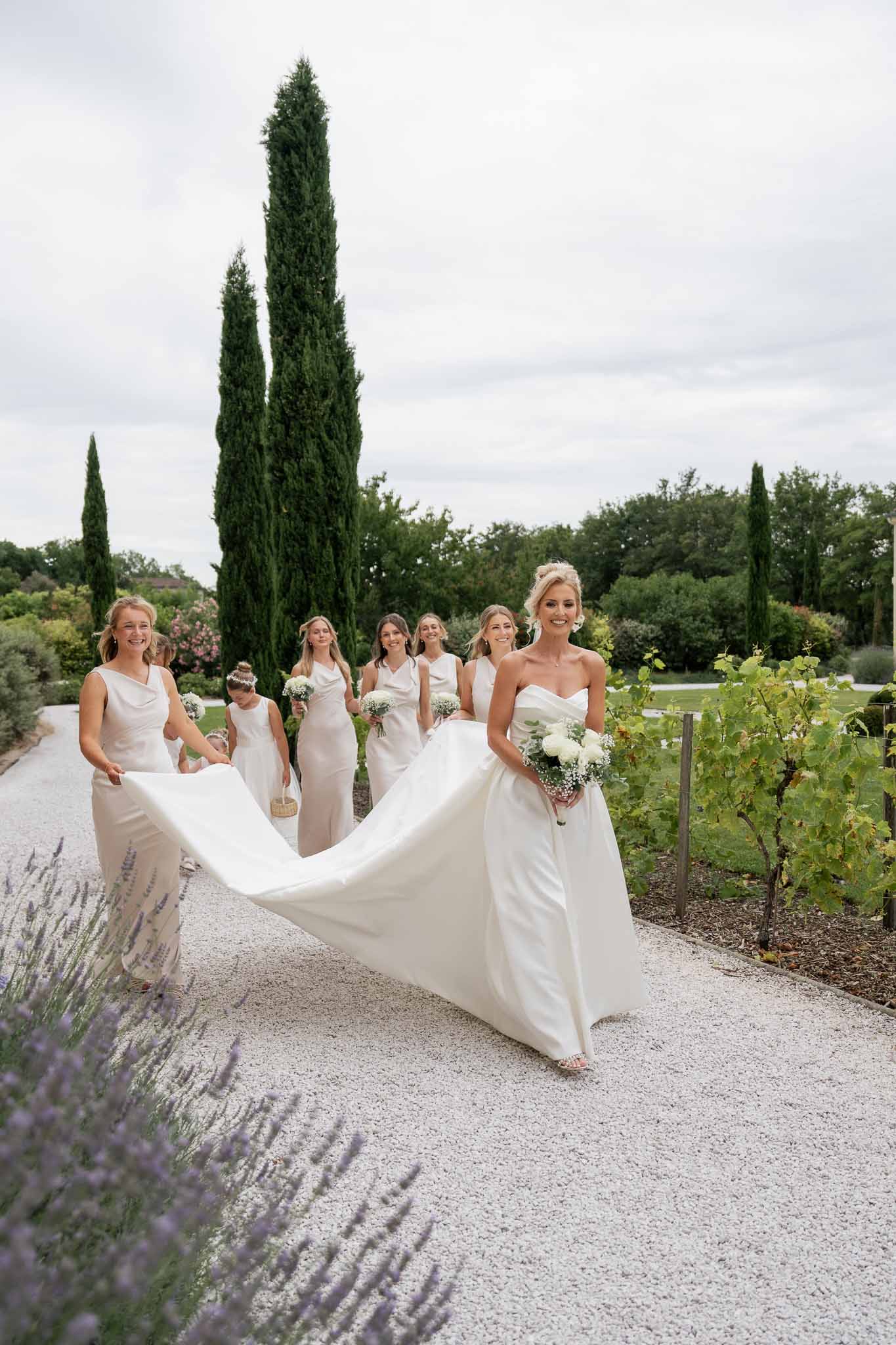 Bride and bridesmaids walking down vineyard pathway with cypress trees and lavender plantings