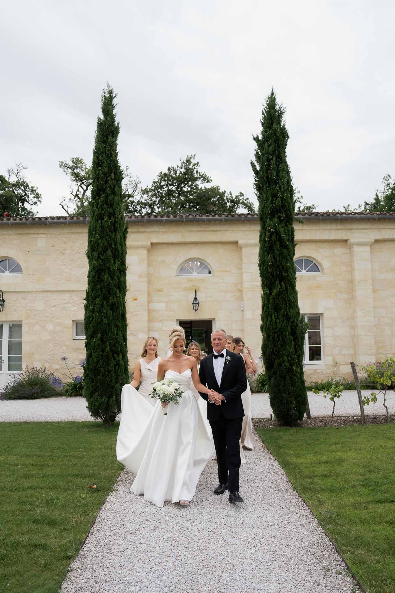 Bride and groom walking down pathway after ceremony at stone château with cypress trees