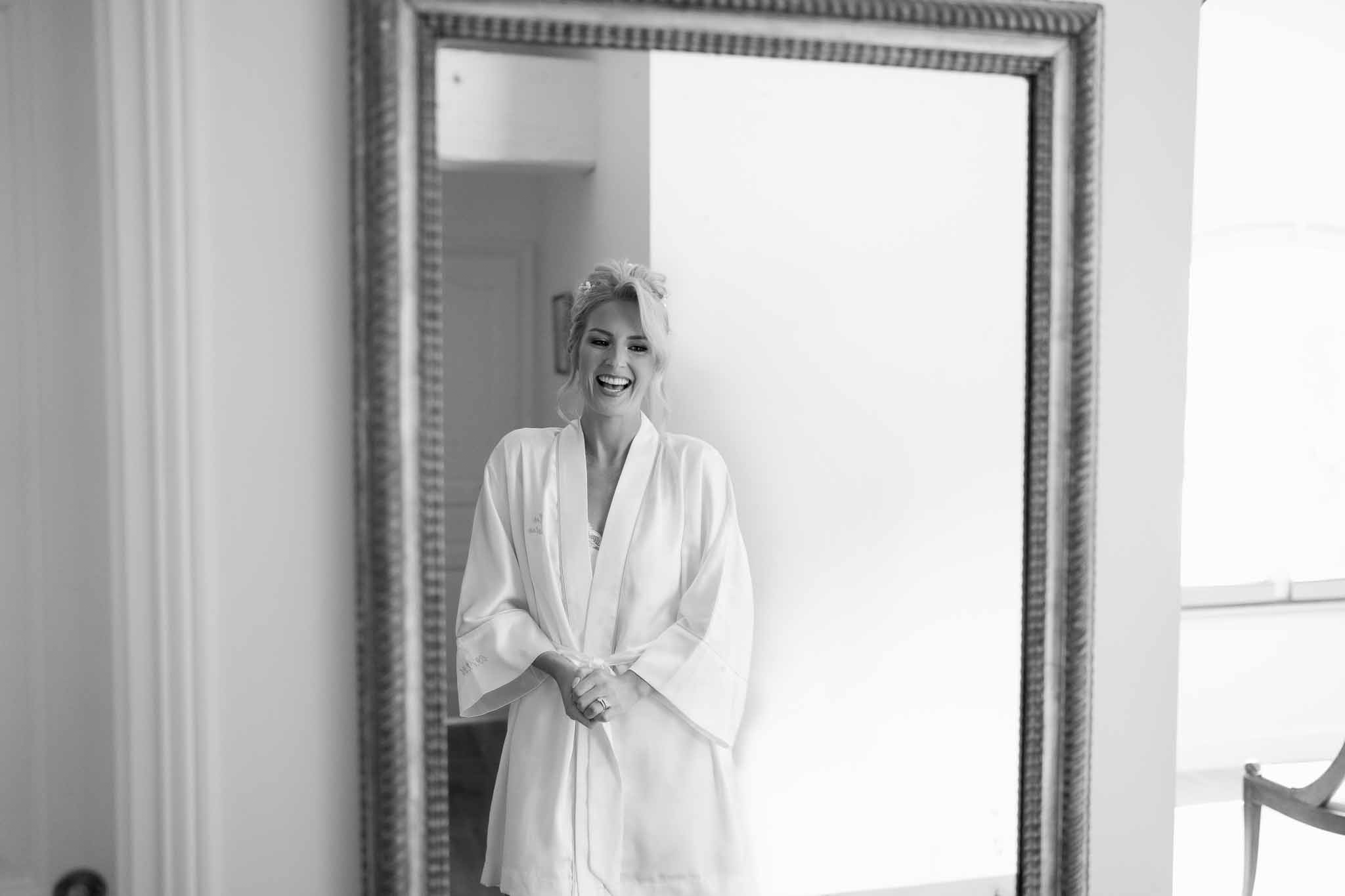 Bride in silk robe smiling during getting ready preparations in bridal suite mirror