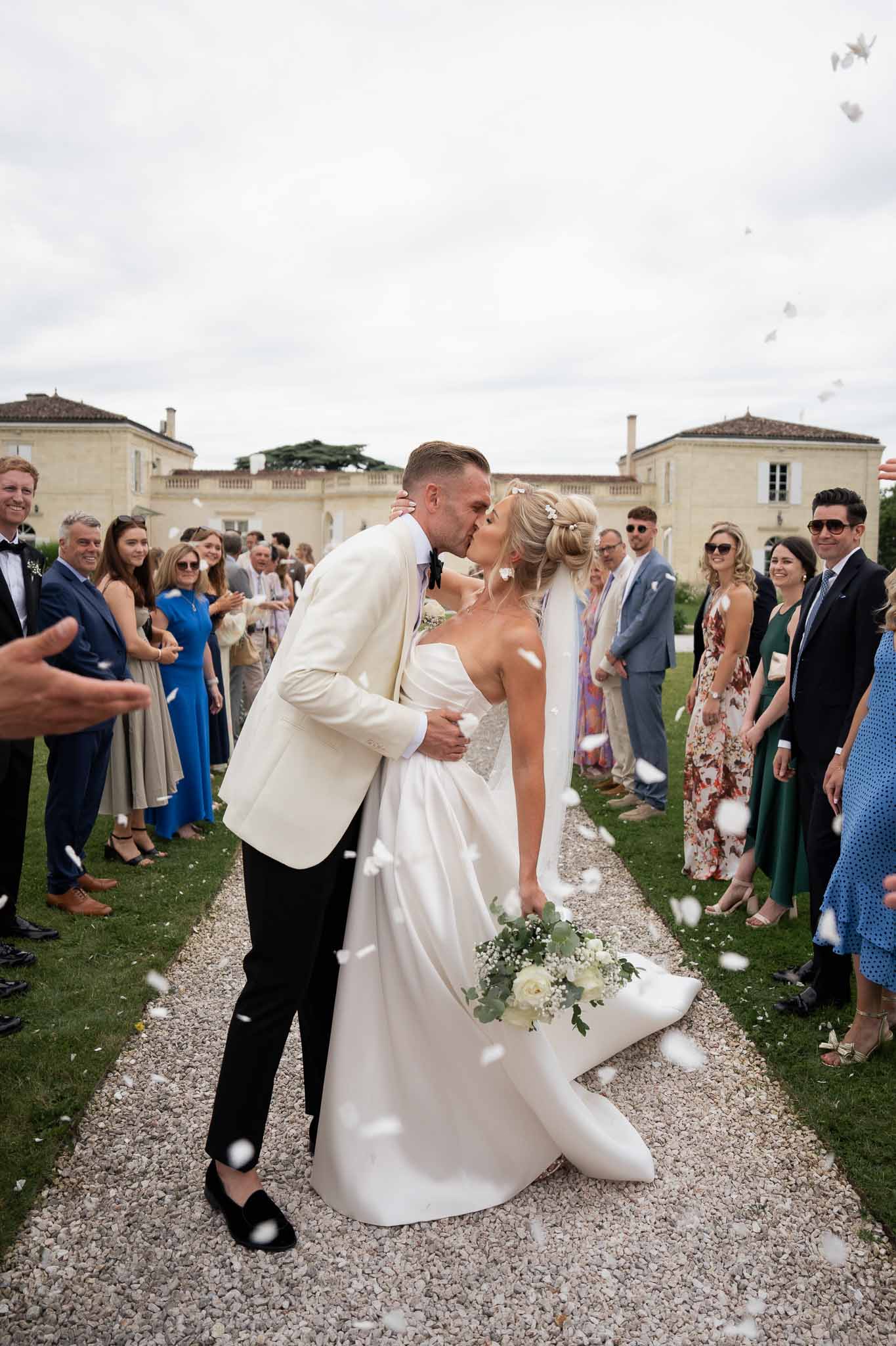 Bride and groom kissing while walking down aisle with applauding guests at French château courtyard