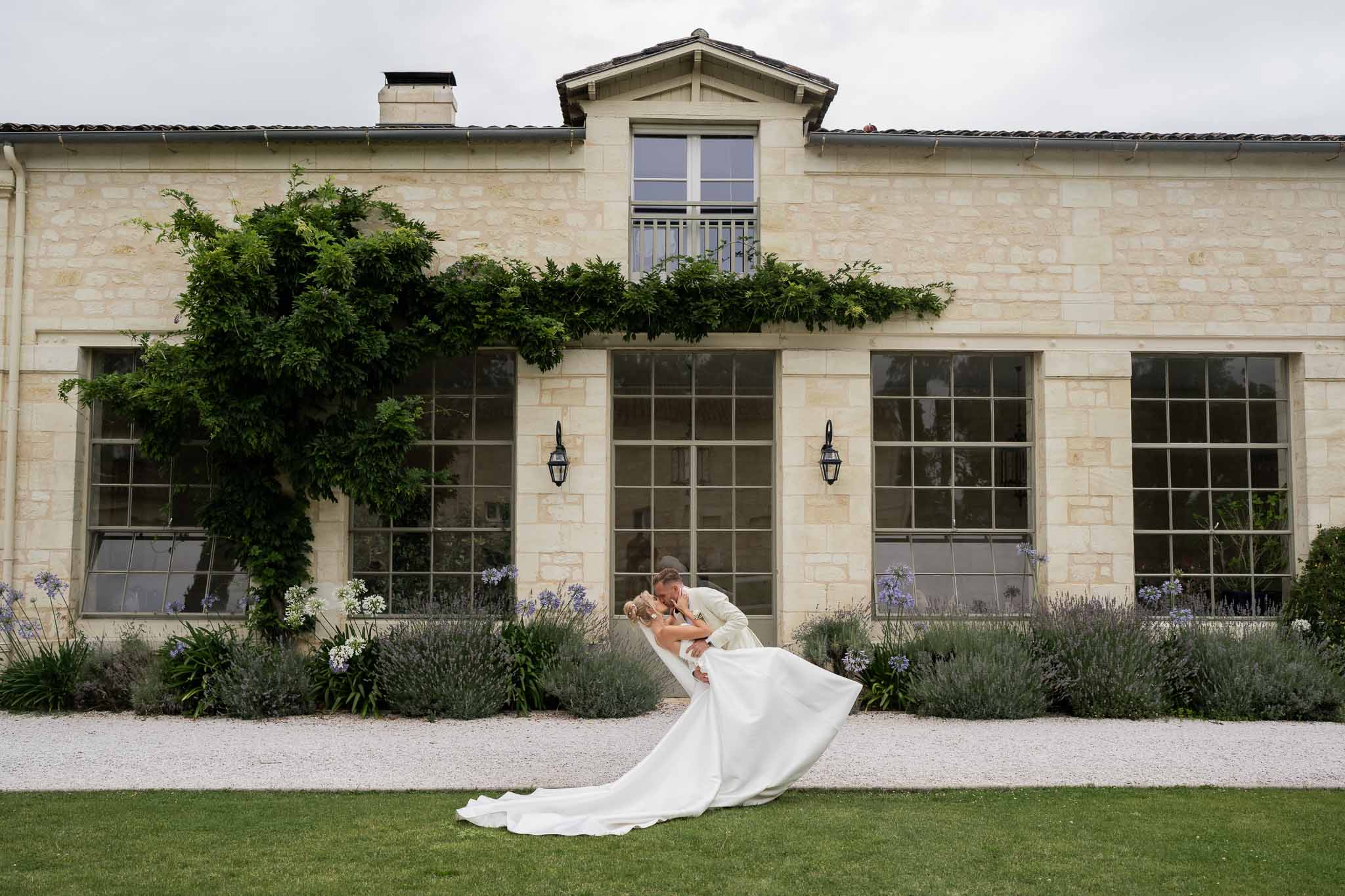 Groom dipping bride in ivory dress on lawn in front of stone estate building with lavender landscaping