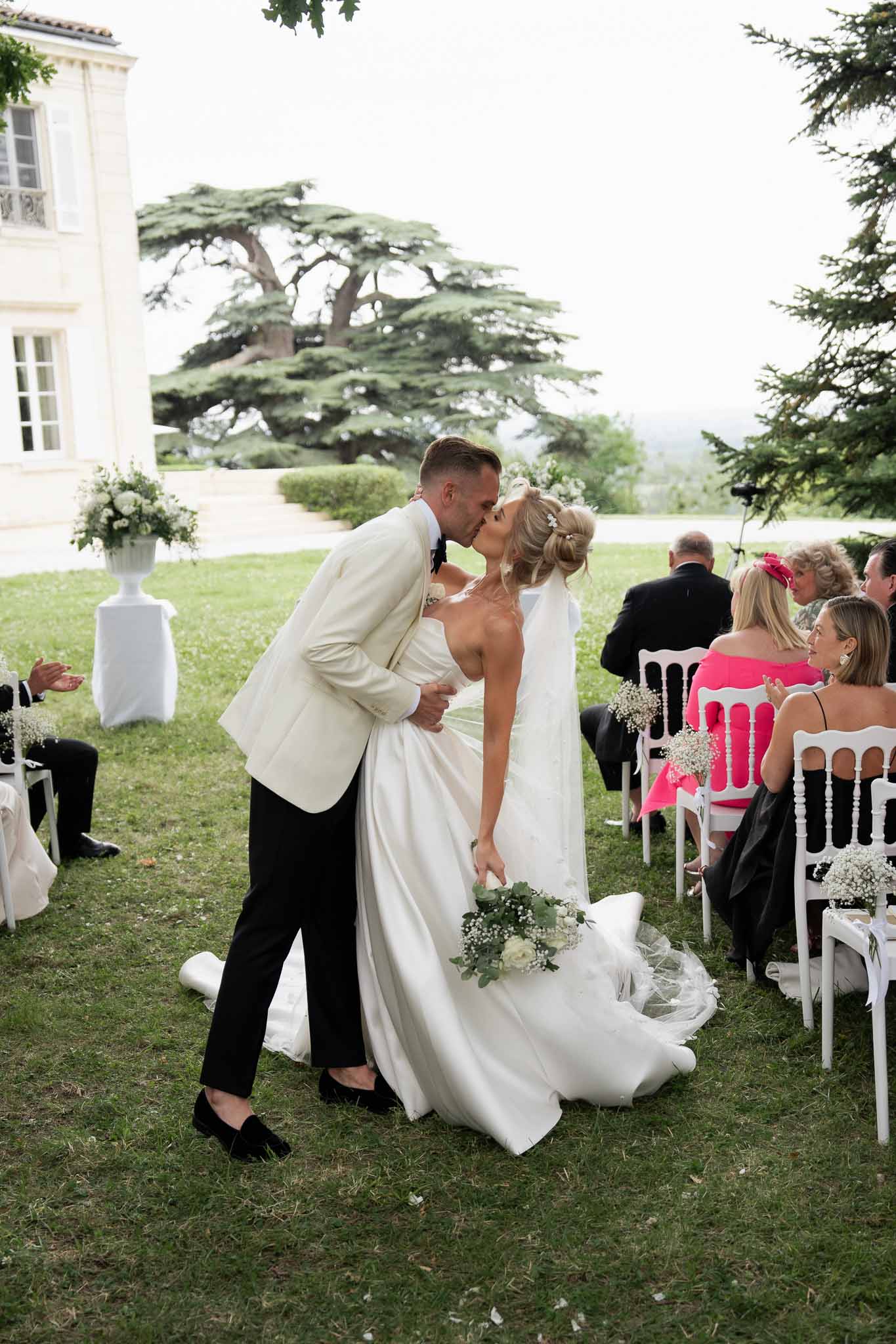 Couple kissing during outdoor wedding ceremony at classic estate with white floral arrangements and cedar tree