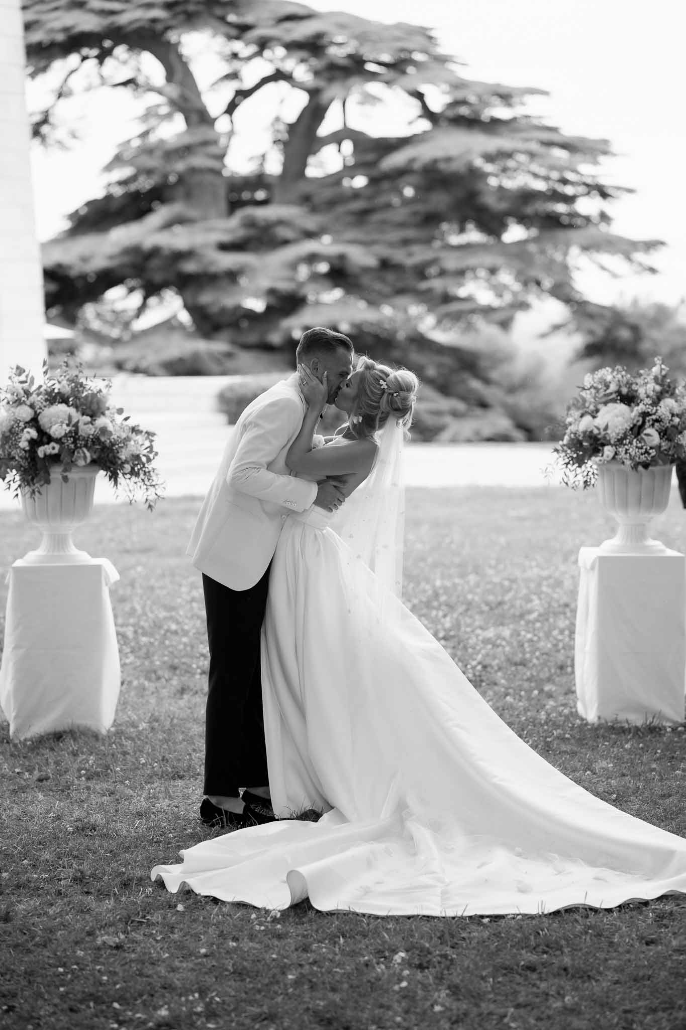 Couple's first kiss after outdoor garden ceremony with white floral arrangements and mature tree backdrop
