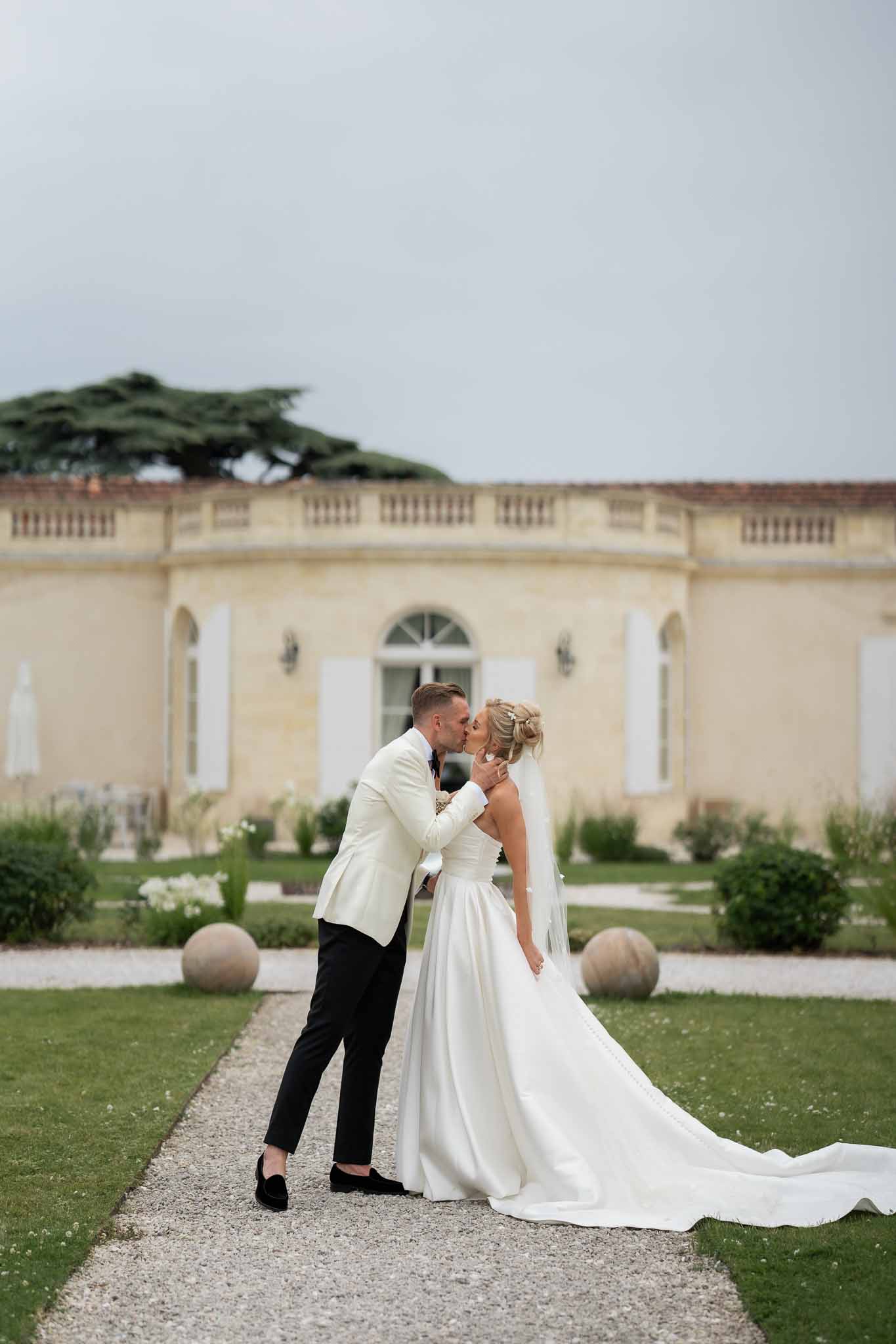 Bride and groom kissing on gravel pathway at classical stone château with formal gardens