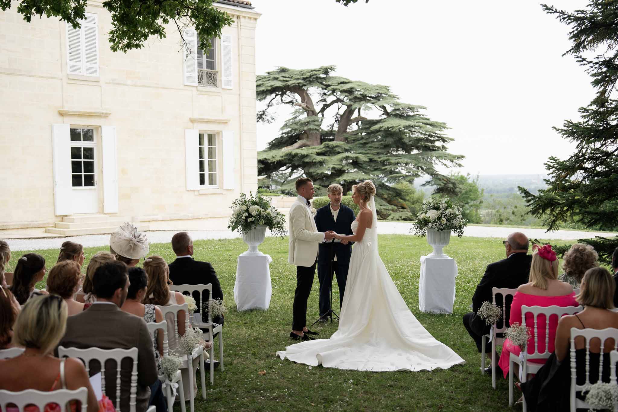 Outdoor wedding ceremony on château lawn with guests in white chairs and stone architecture backdrop