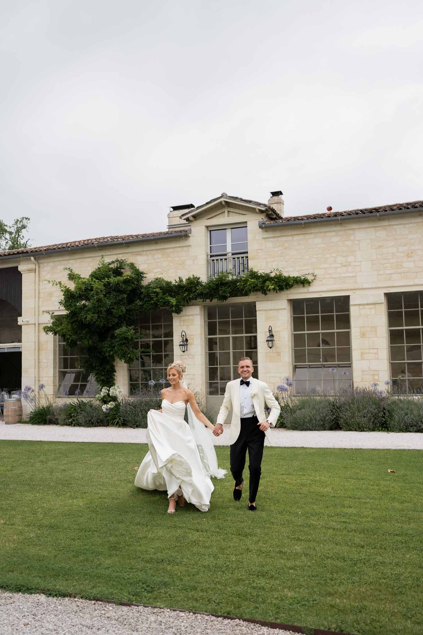 Bride and groom running across lawn in front of classical stone estate building