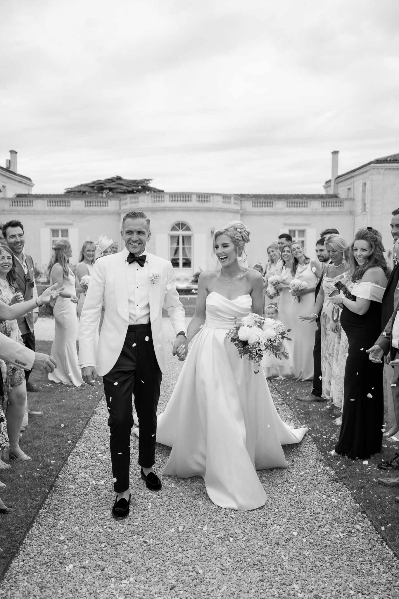 Newlyweds walking down gravel path after ceremony surrounded by guests in classical stone courtyard