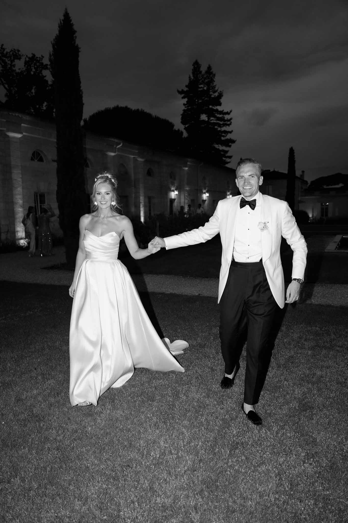 Bride and groom walking hand-in-hand across illuminated courtyard with classical architecture