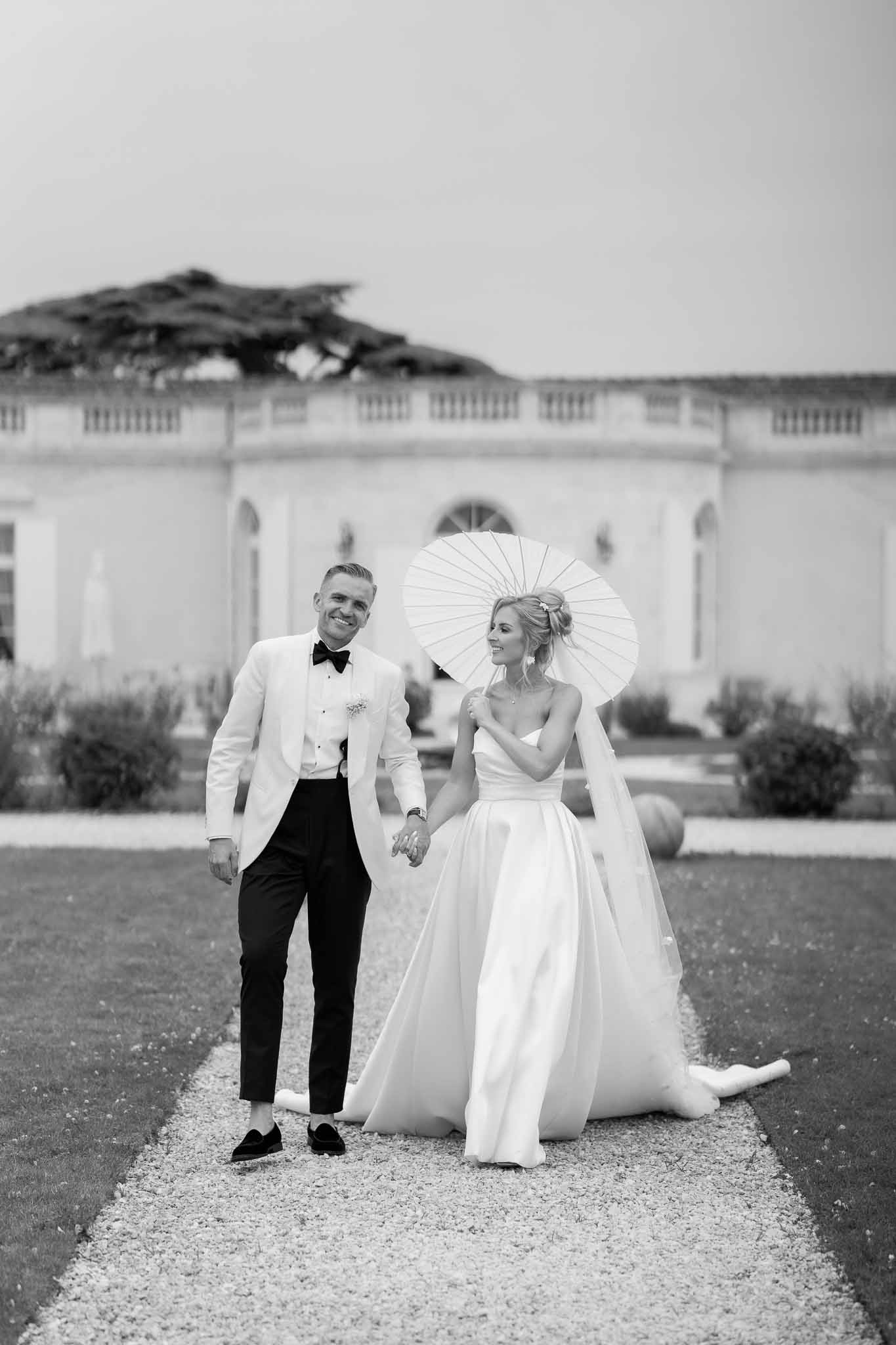 Newlyweds walking hand-in-hand in front of neoclassical building with columns and formal gardens