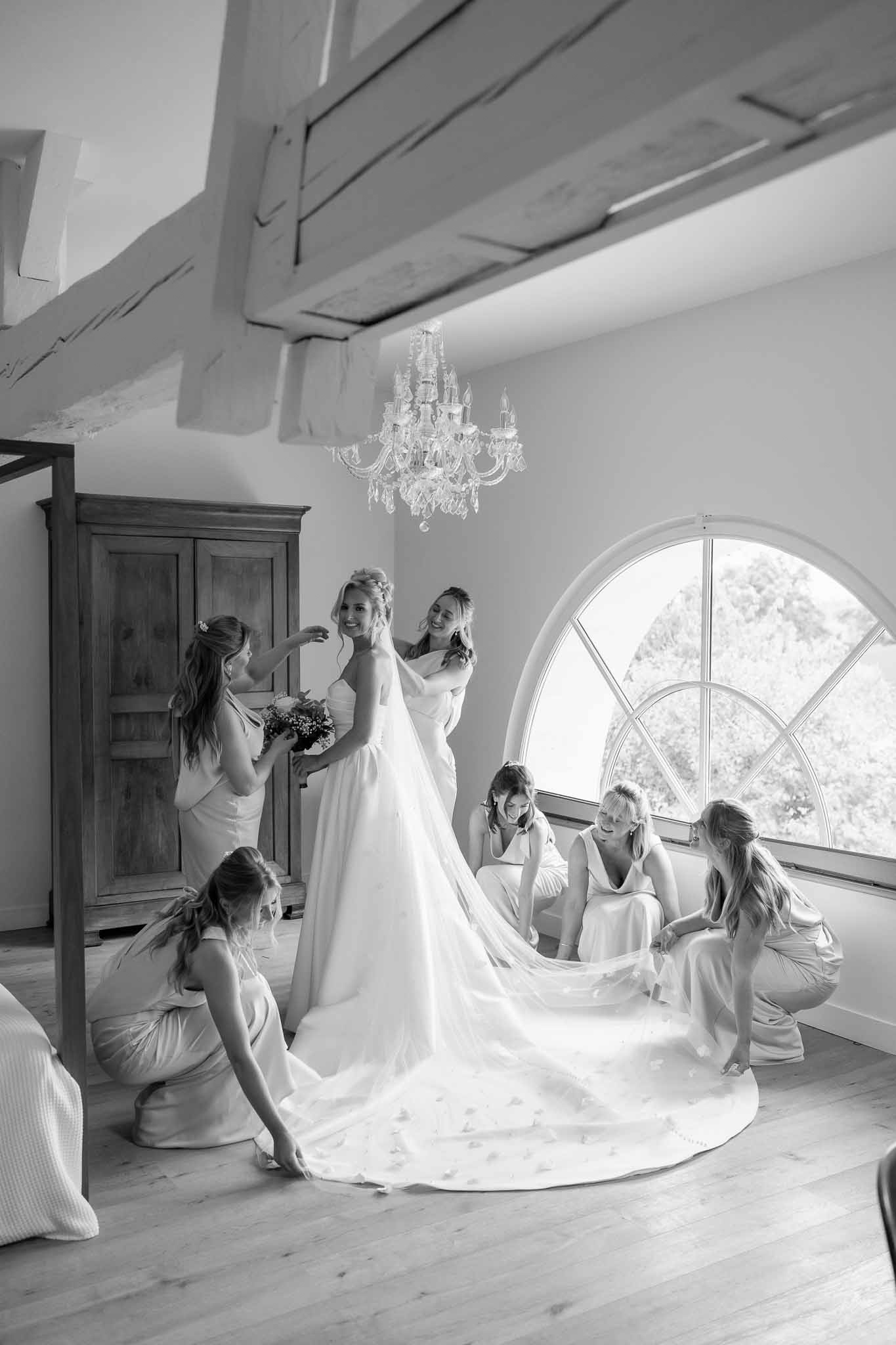 Bride in white gown surrounded by bridesmaids getting ready in modern interior space with arched window