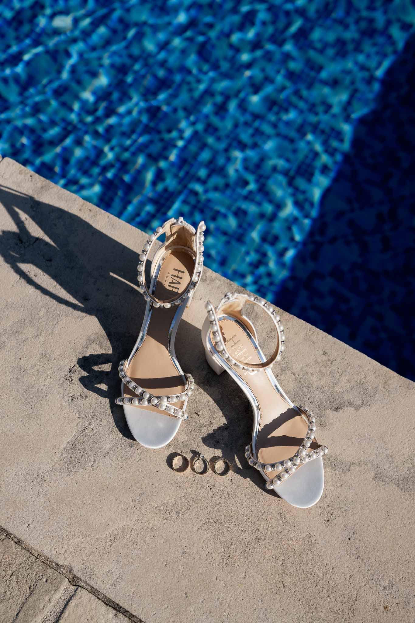 Bride's white pearl-embellished sandals and wedding rings arranged on pool deck at poolside wedding venue