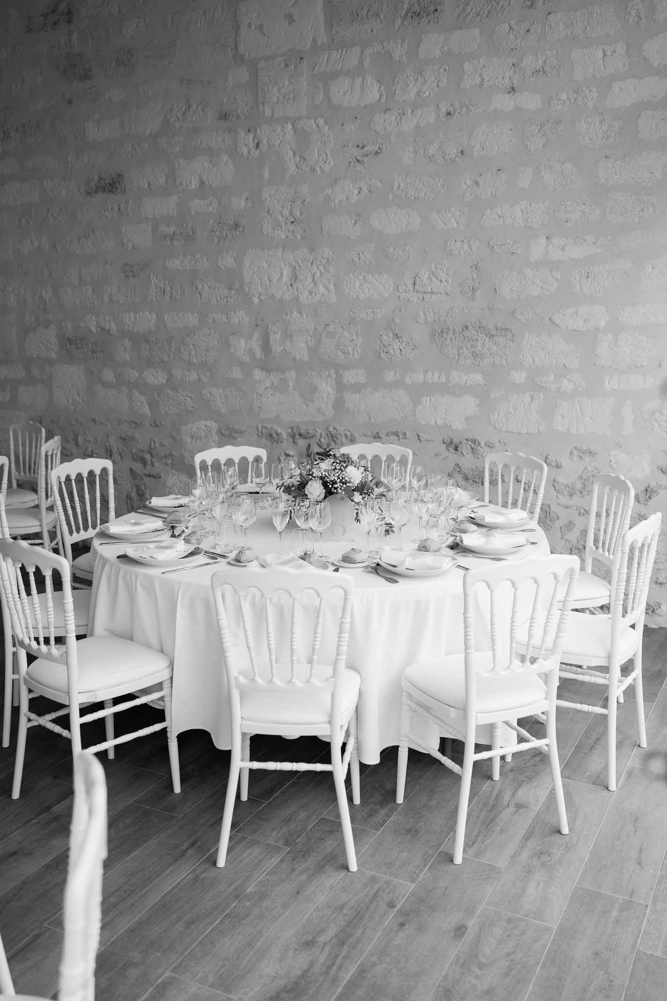Round reception table setting with white linens and floral centerpiece against whitewashed brick wall
