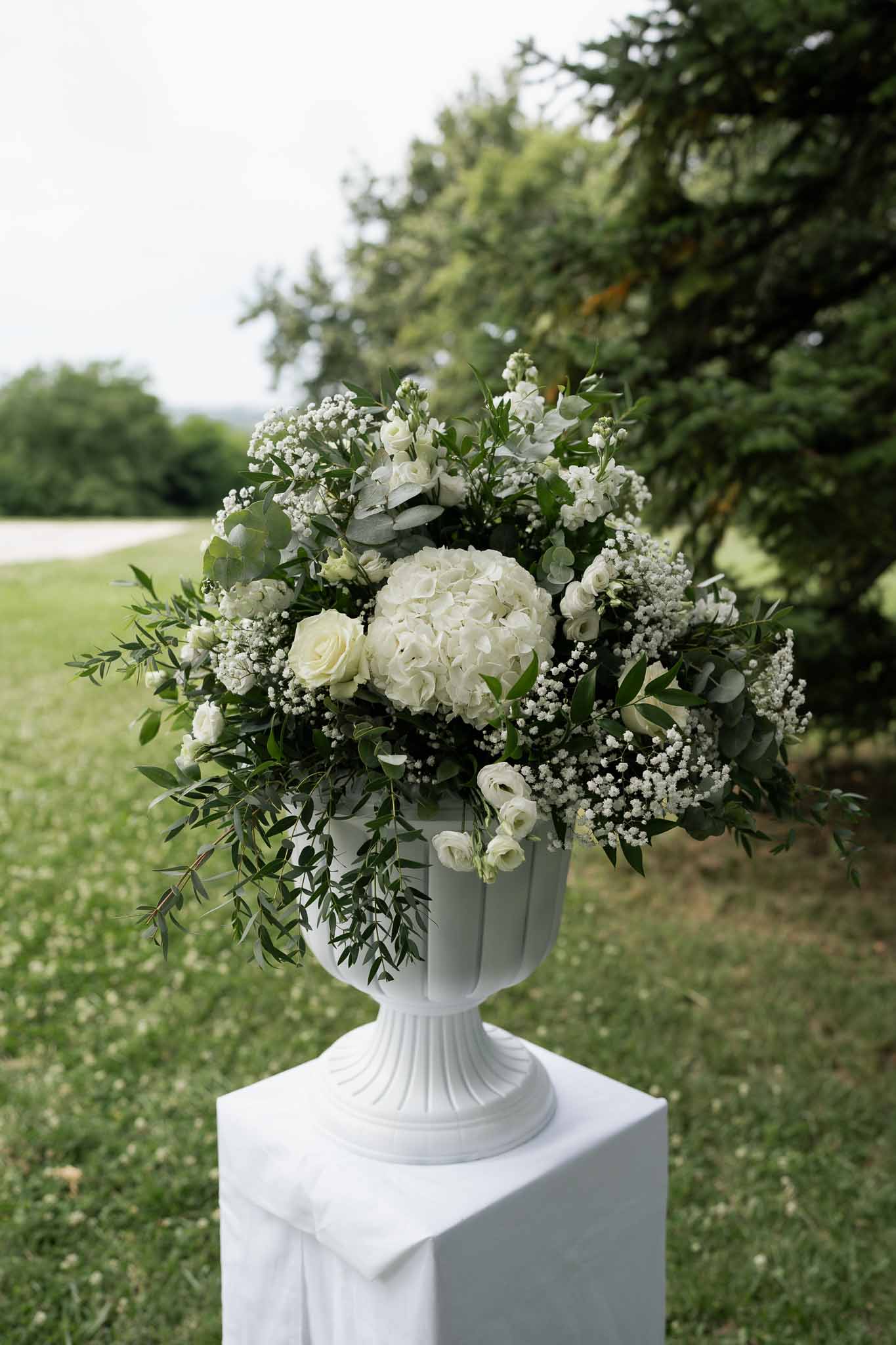 Ivory hydrangea and rose floral arrangement on white pedestal urn at outdoor garden wedding reception
