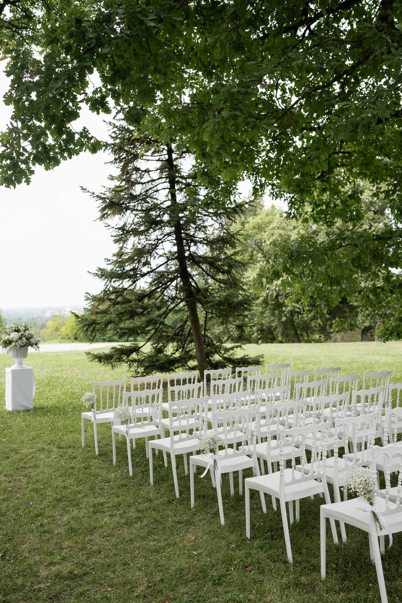Outdoor wedding ceremony setup with white chairs on lakeside garden lawn with trees overhead
