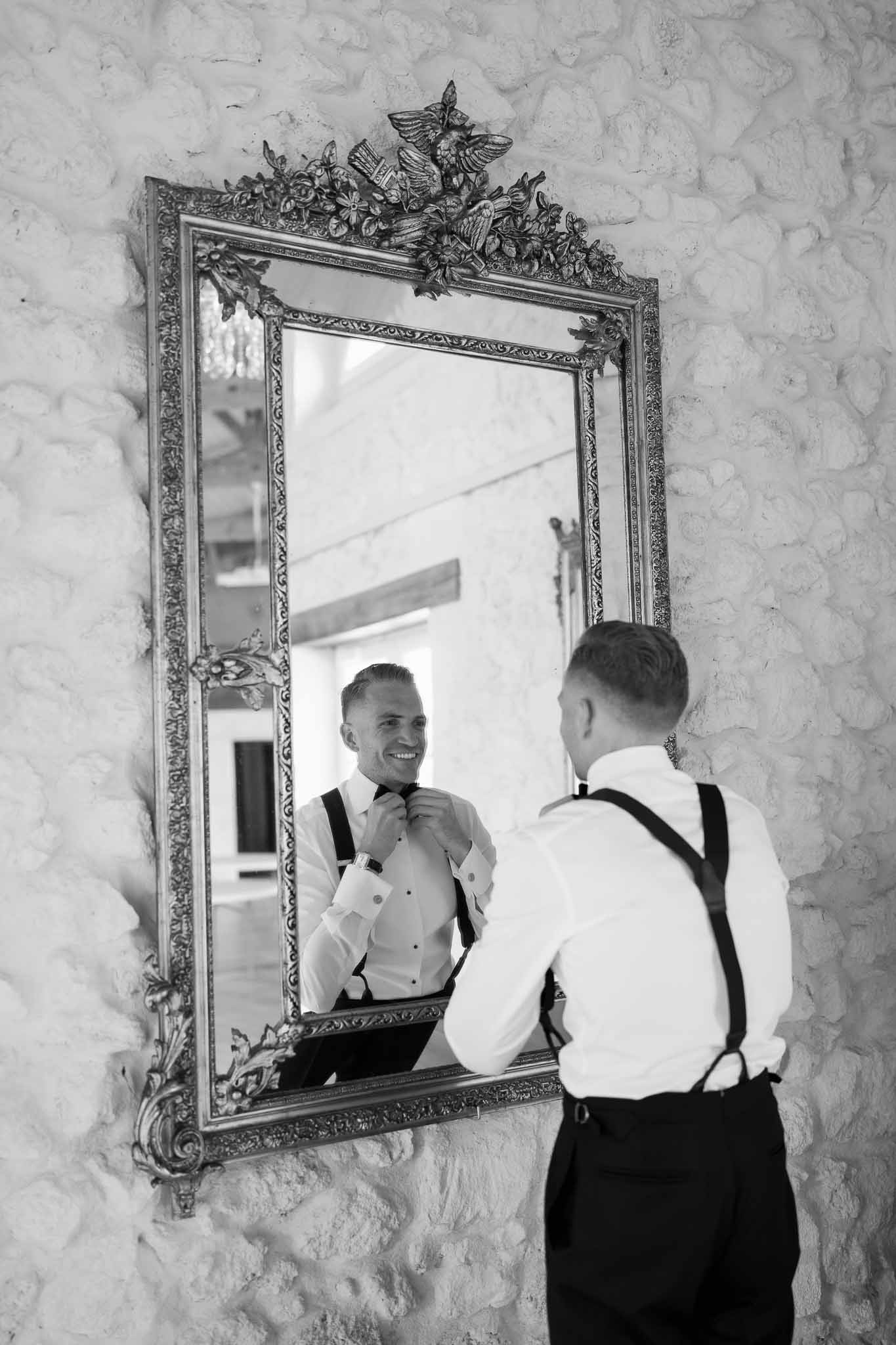 Groom adjusting bow tie in ornate mirror during wedding preparation