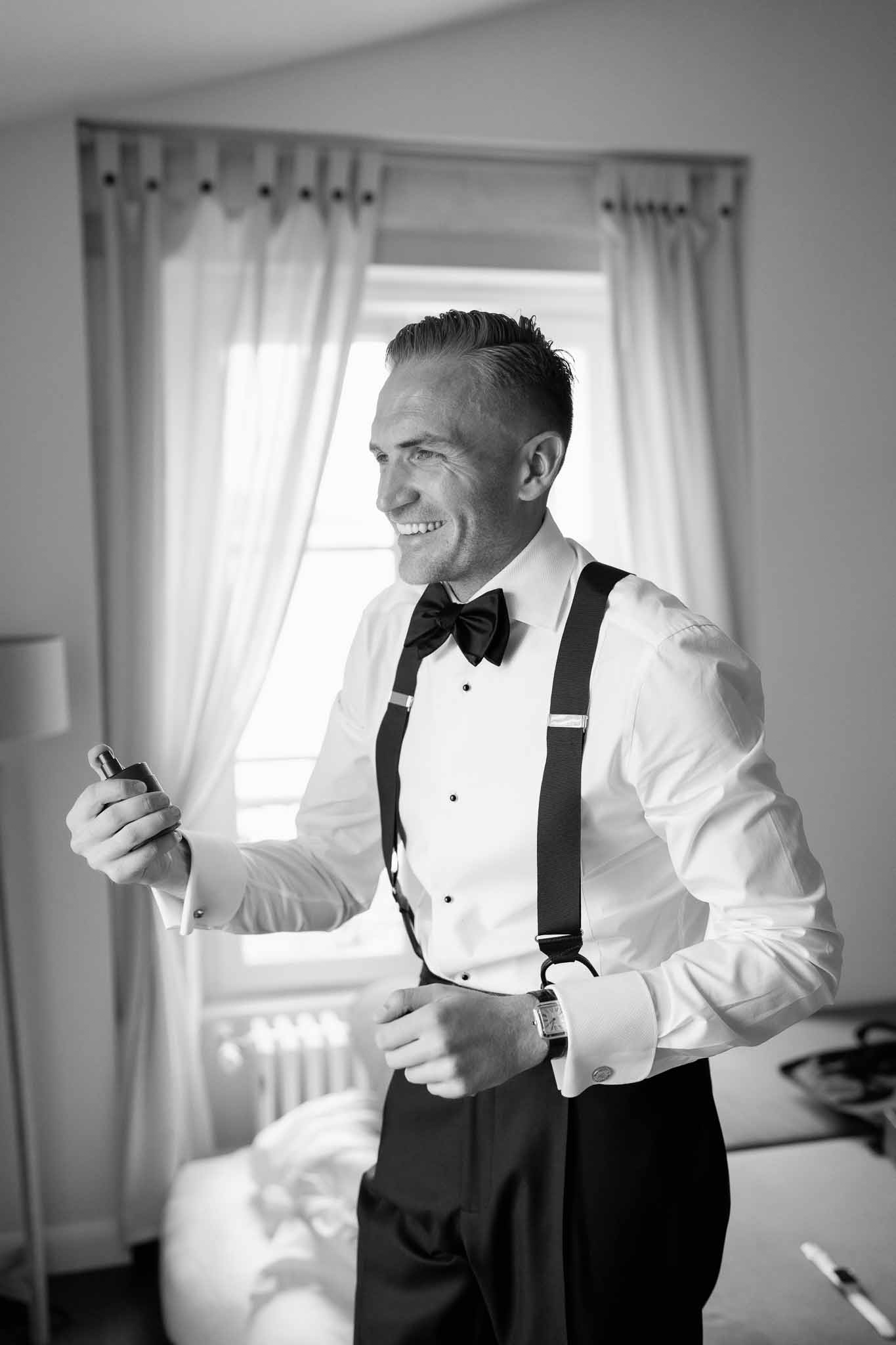 Groom in formal evening wear with bow tie and suspenders holding cufflinks during getting ready in bedroom