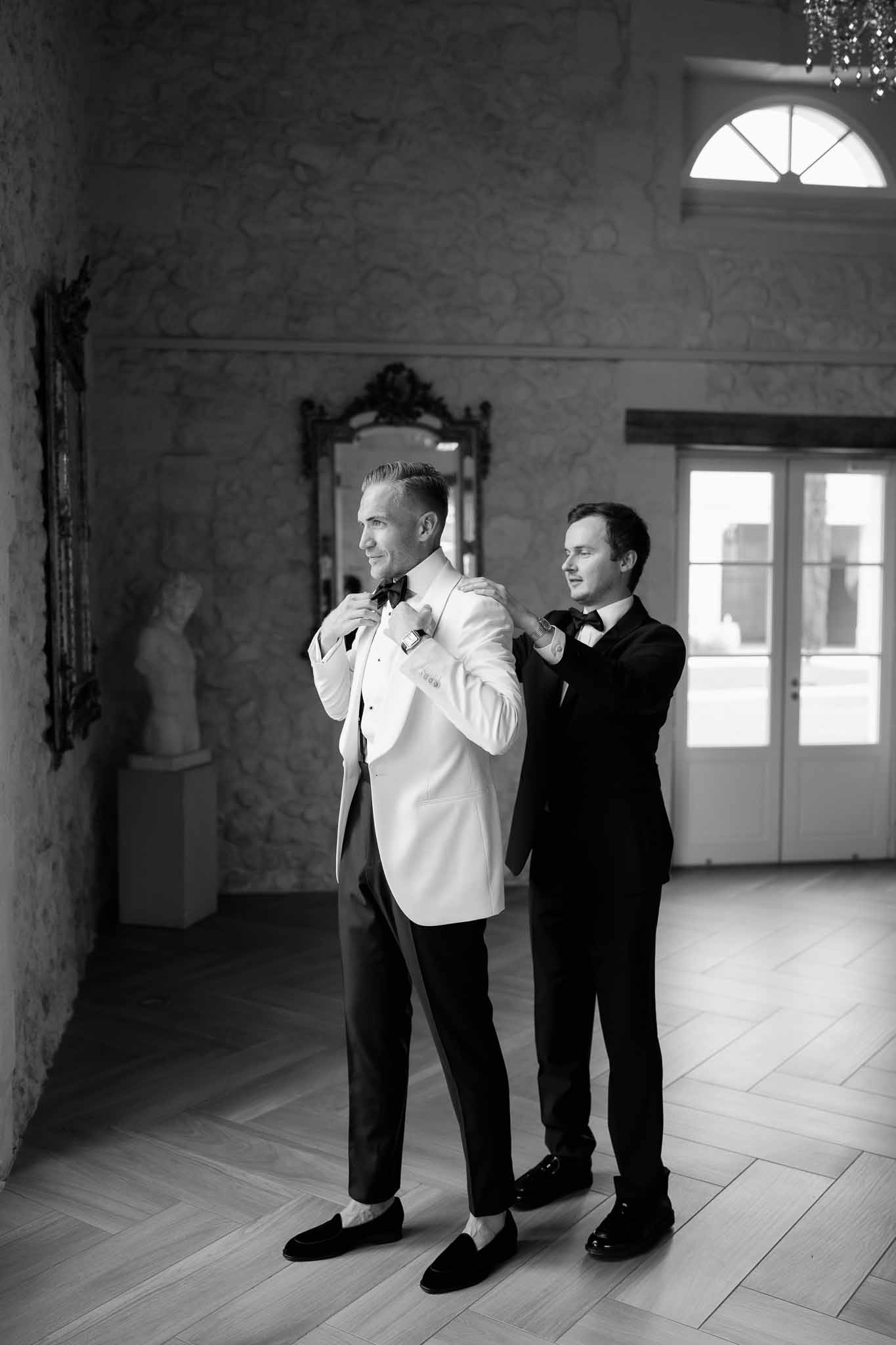 Groom adjusting white dinner jacket with groomsman assistance in elegant interior hallway