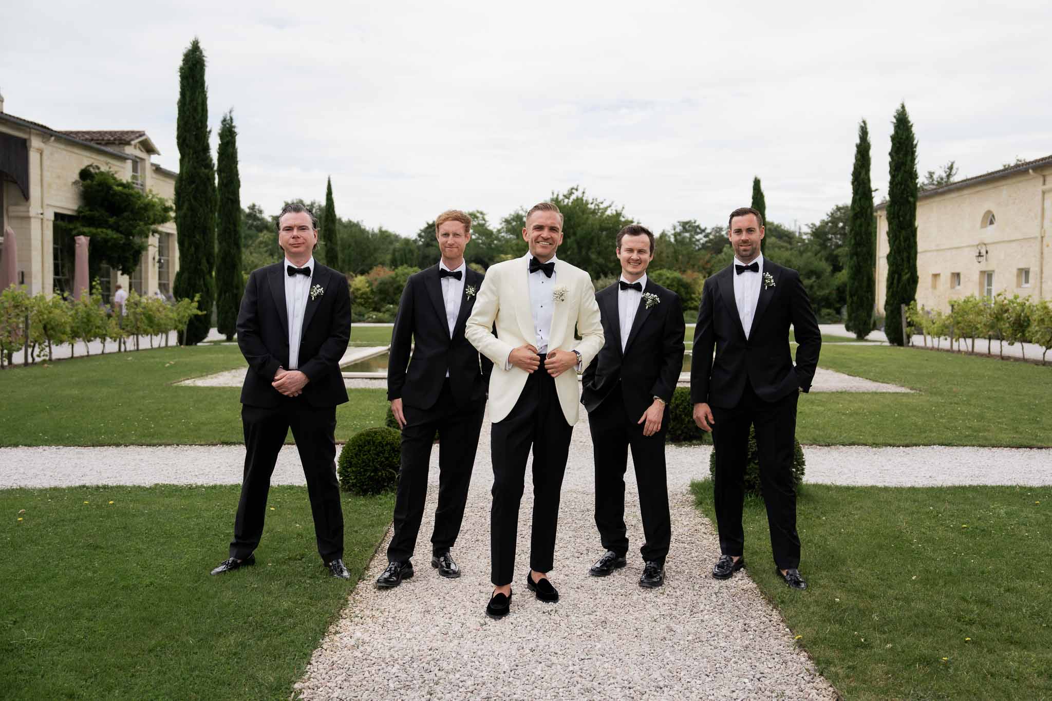 Groom in cream jacket with groomsmen in black tuxedos on estate garden pathway