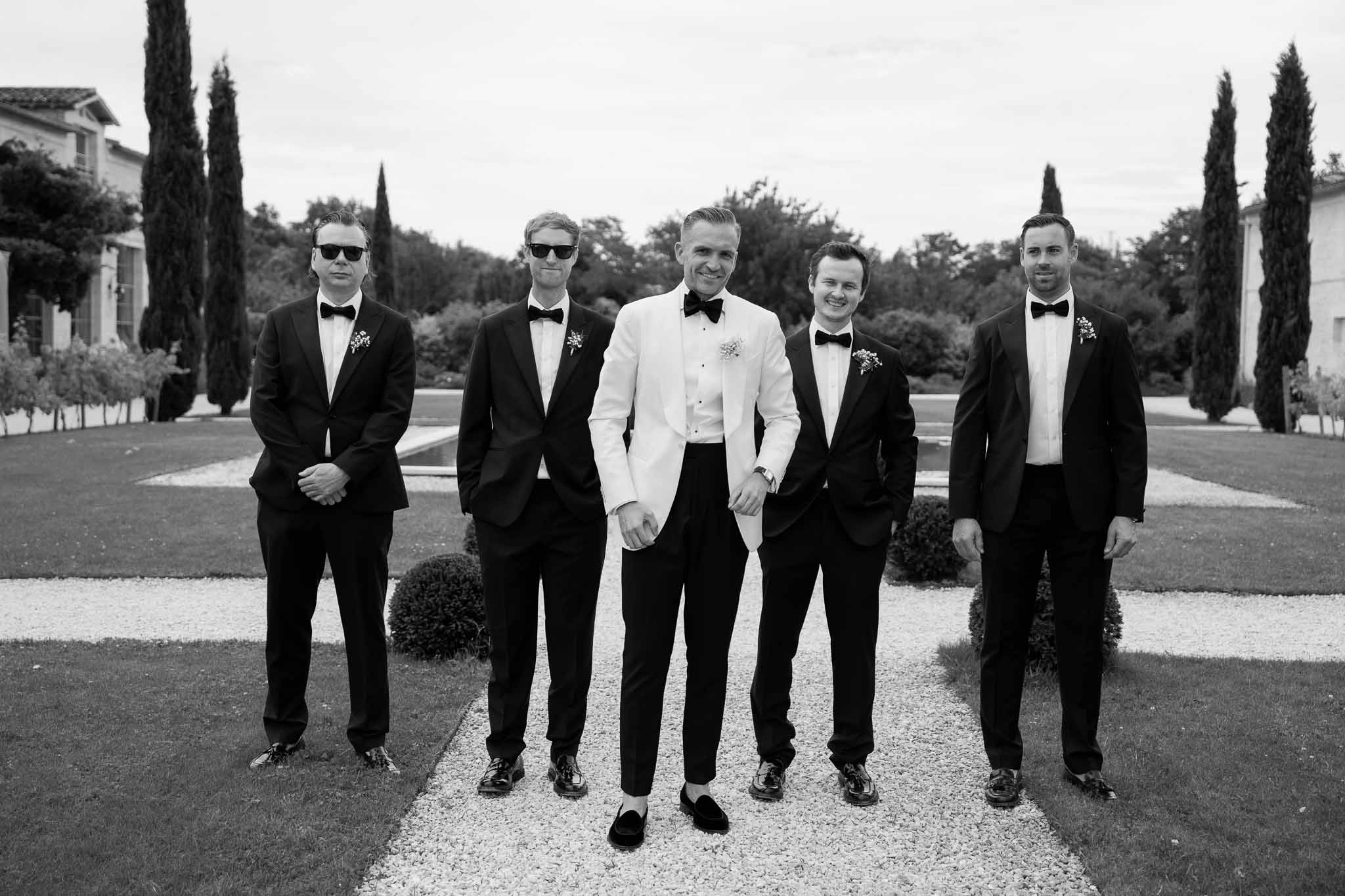 Groom and groomsmen in formal attire standing on estate grounds with cypress trees and manicured gardens
