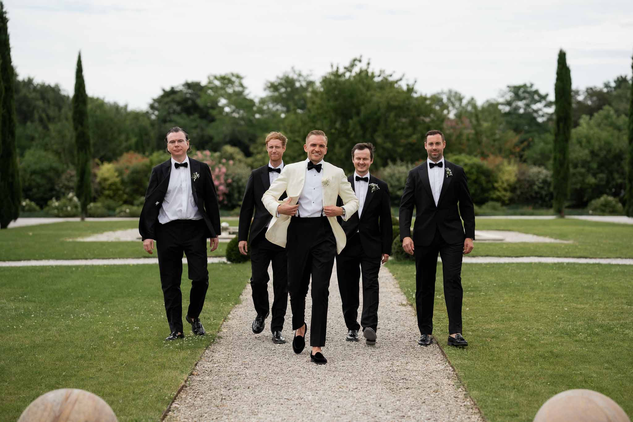 Groom and groomsmen walking through formal garden pathway at wedding venue