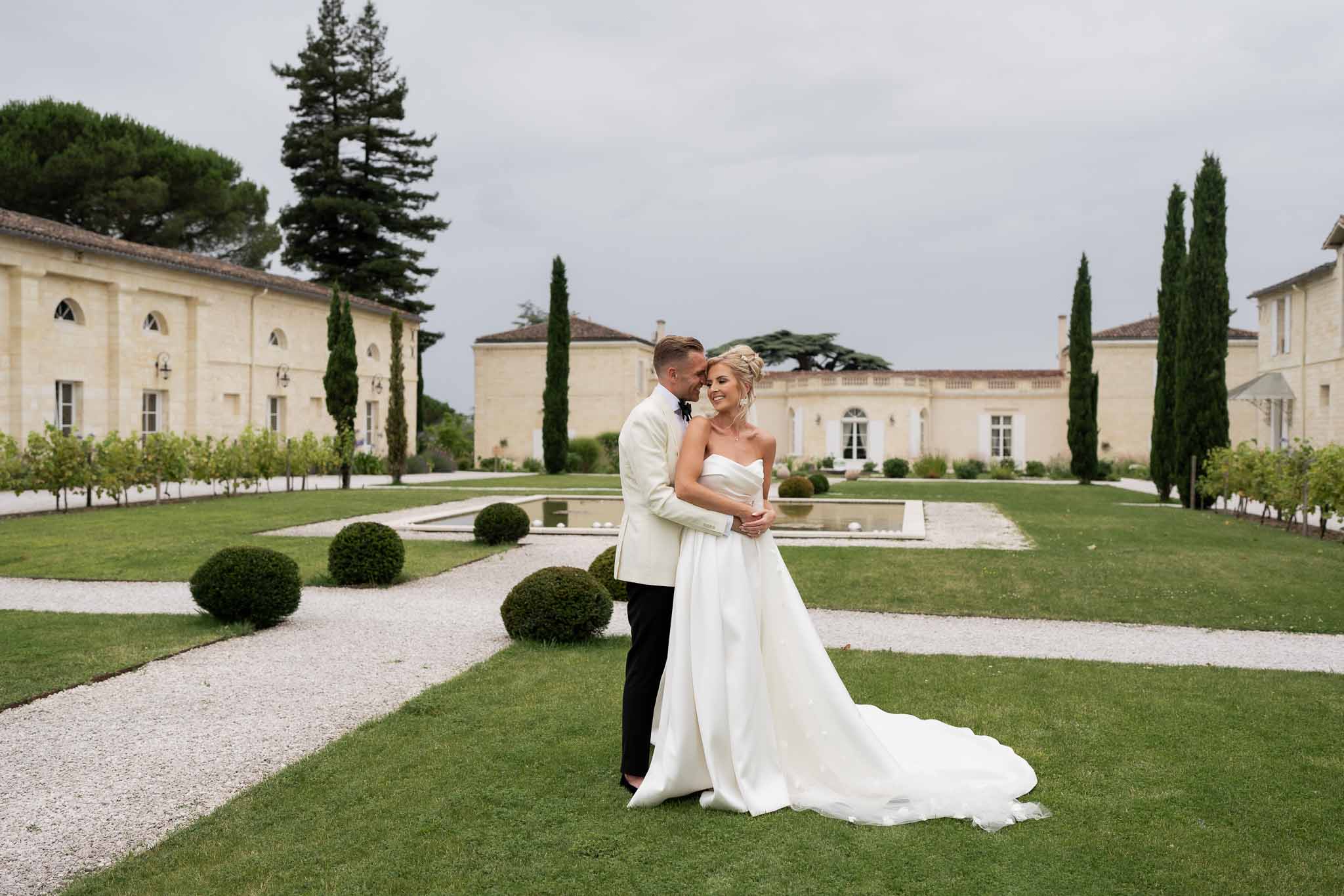 Bride and groom posing together in manicured grounds of classical stone estate with reflecting pool