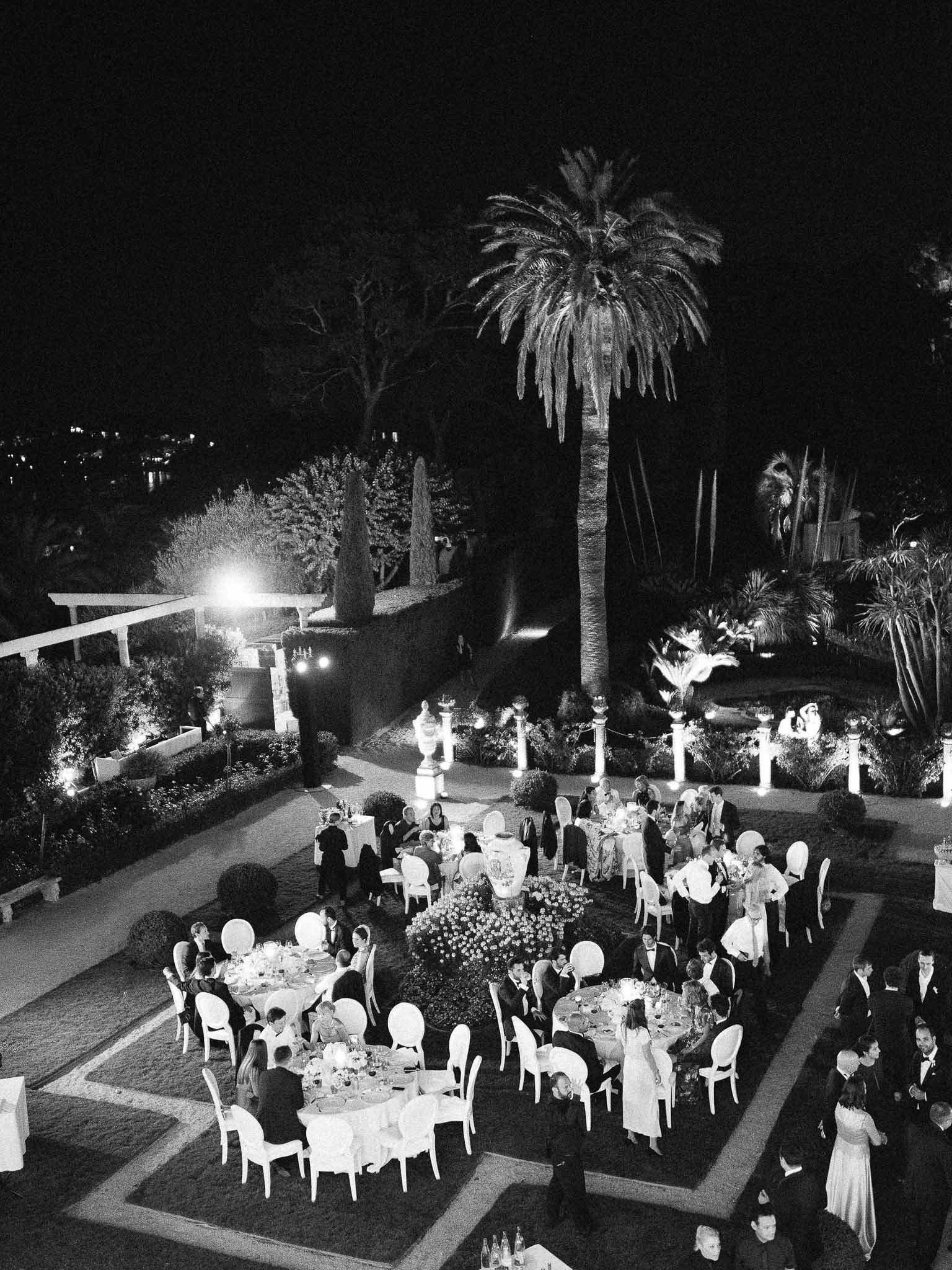 Aerial view of nighttime wedding reception with guests dining at round tables in garden courtyard venue