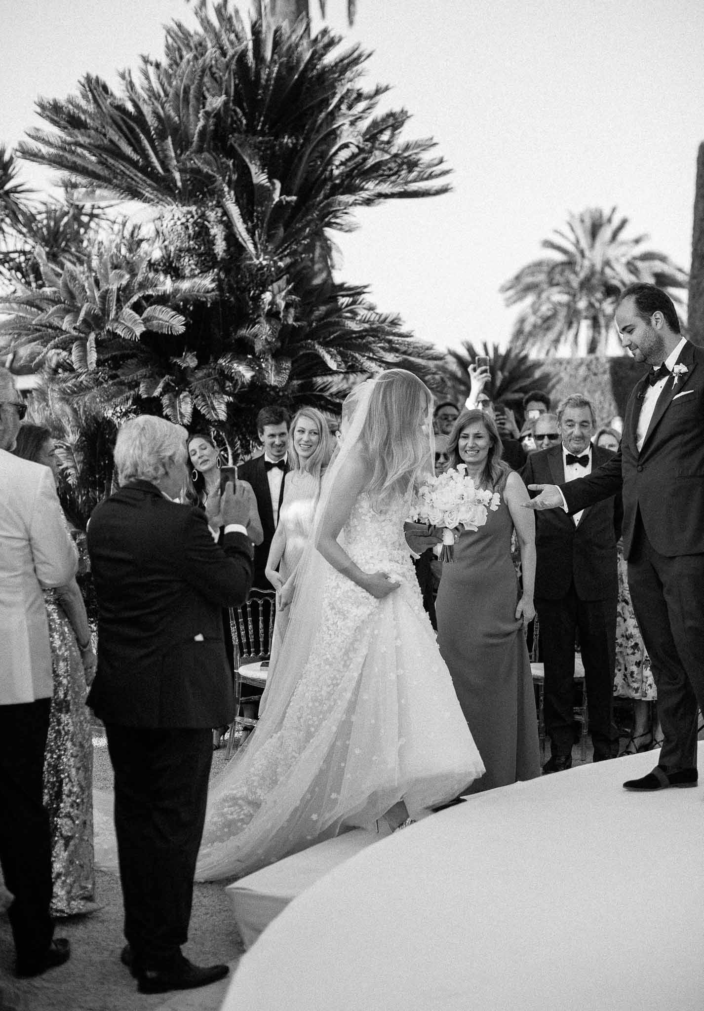 Bride and groom during outdoor wedding ceremony with palm trees and formal guests