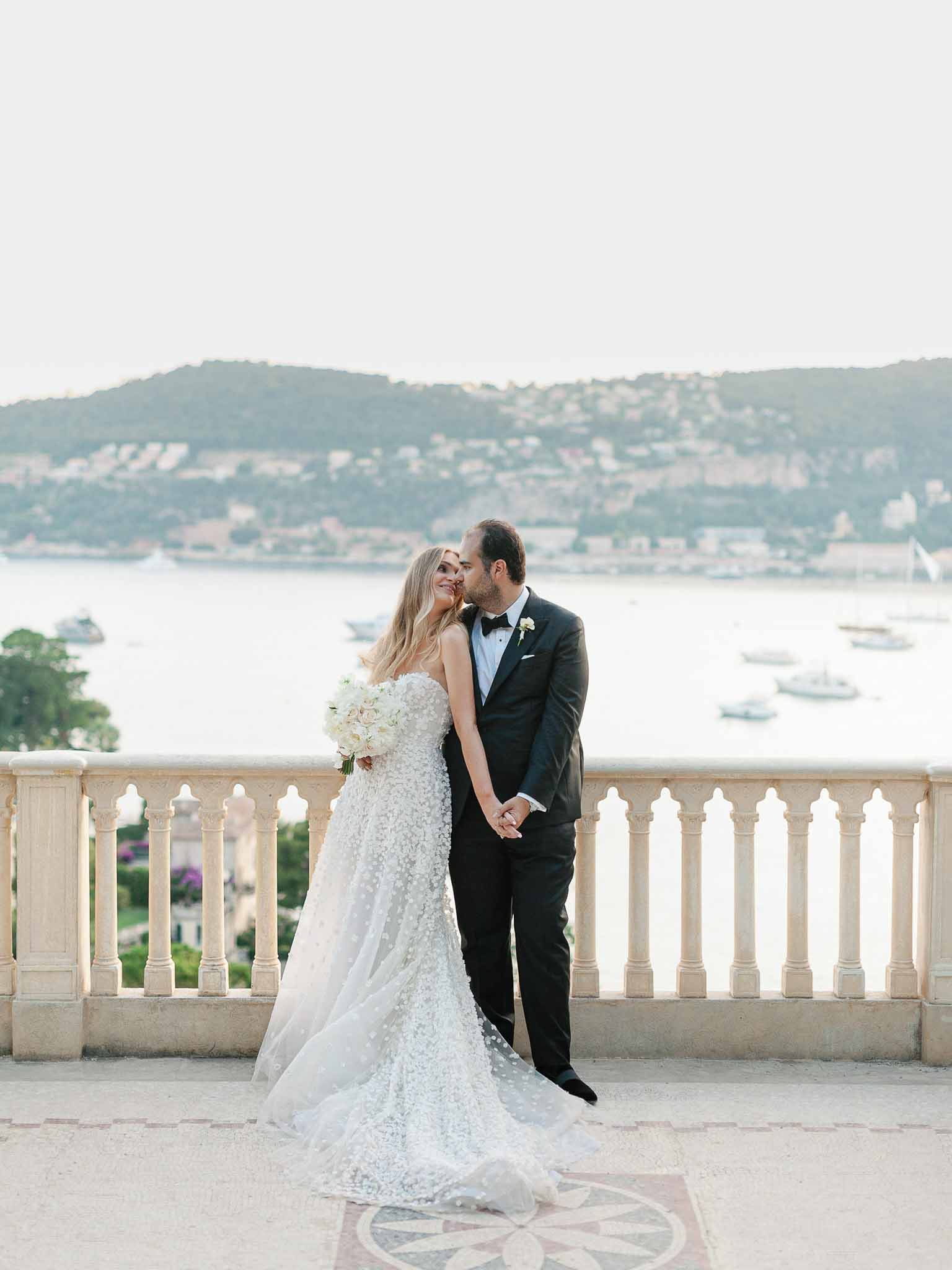 Bride and groom portrait on stone terrace overlooking waterfront bay with boats and hills
