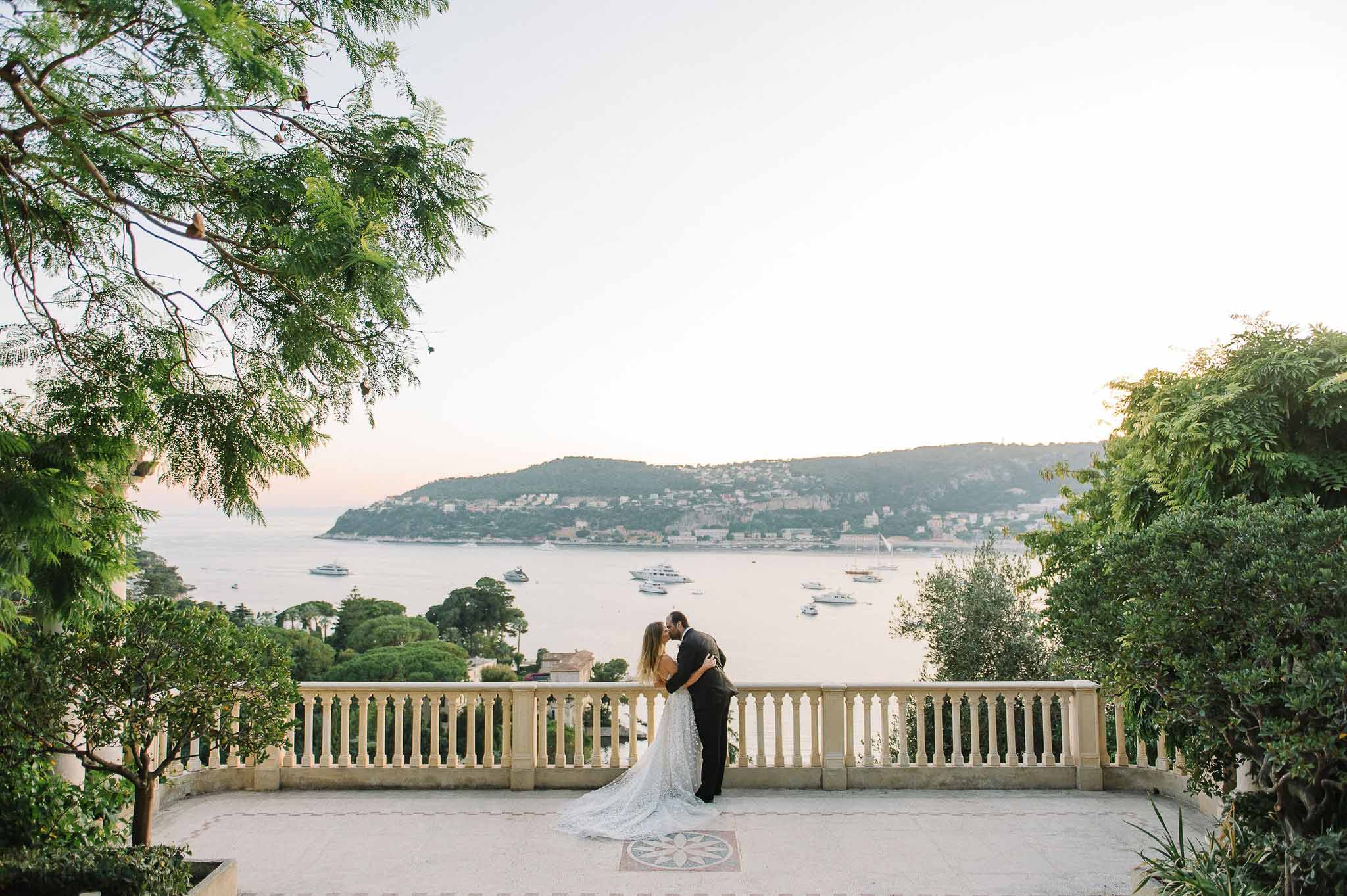 Bride and groom kissing on Mediterranean terrace overlooking bay with sailboats