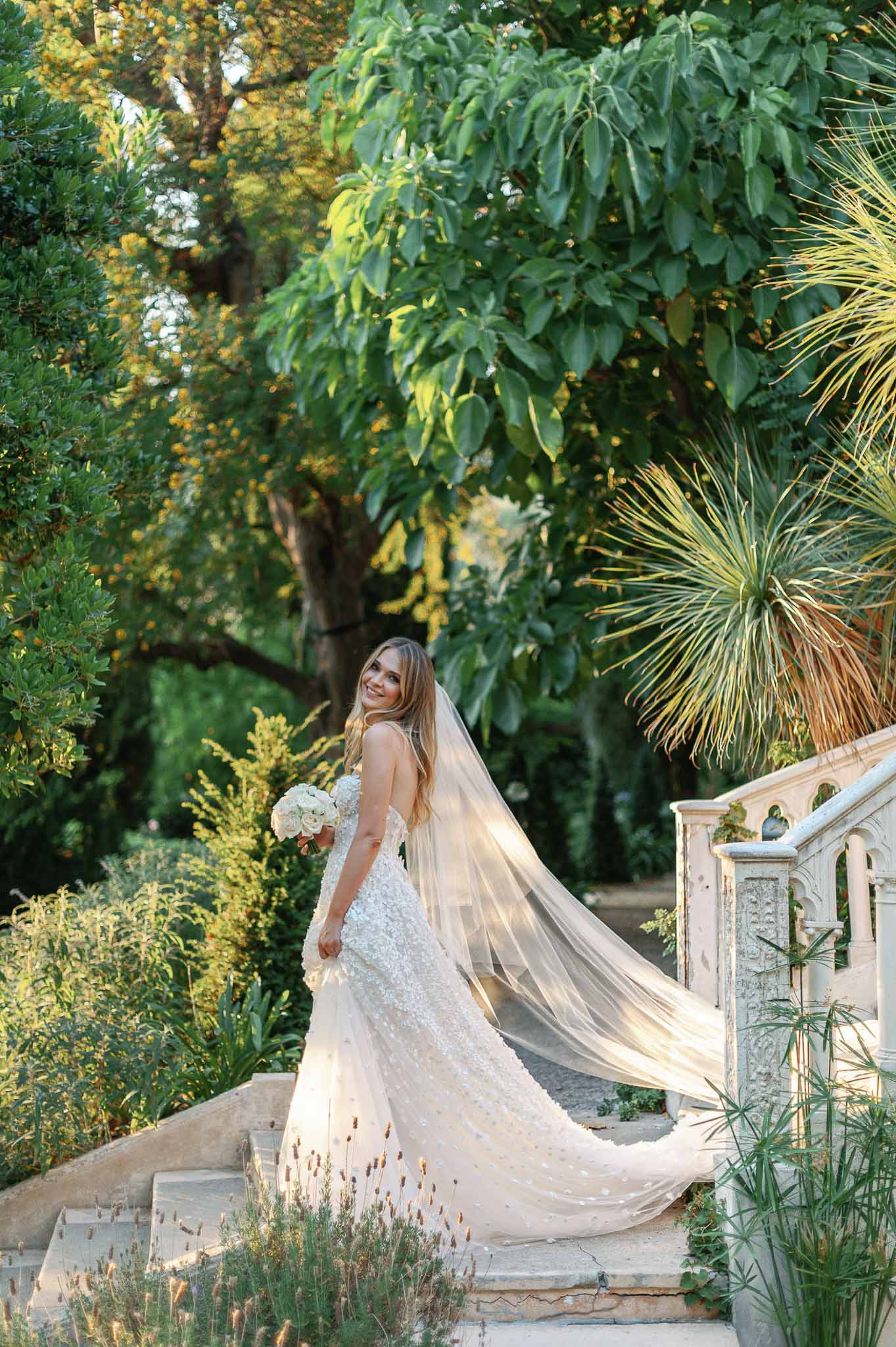 Bride in beaded ivory gown holding white bouquet in classical garden setting with stone pathway