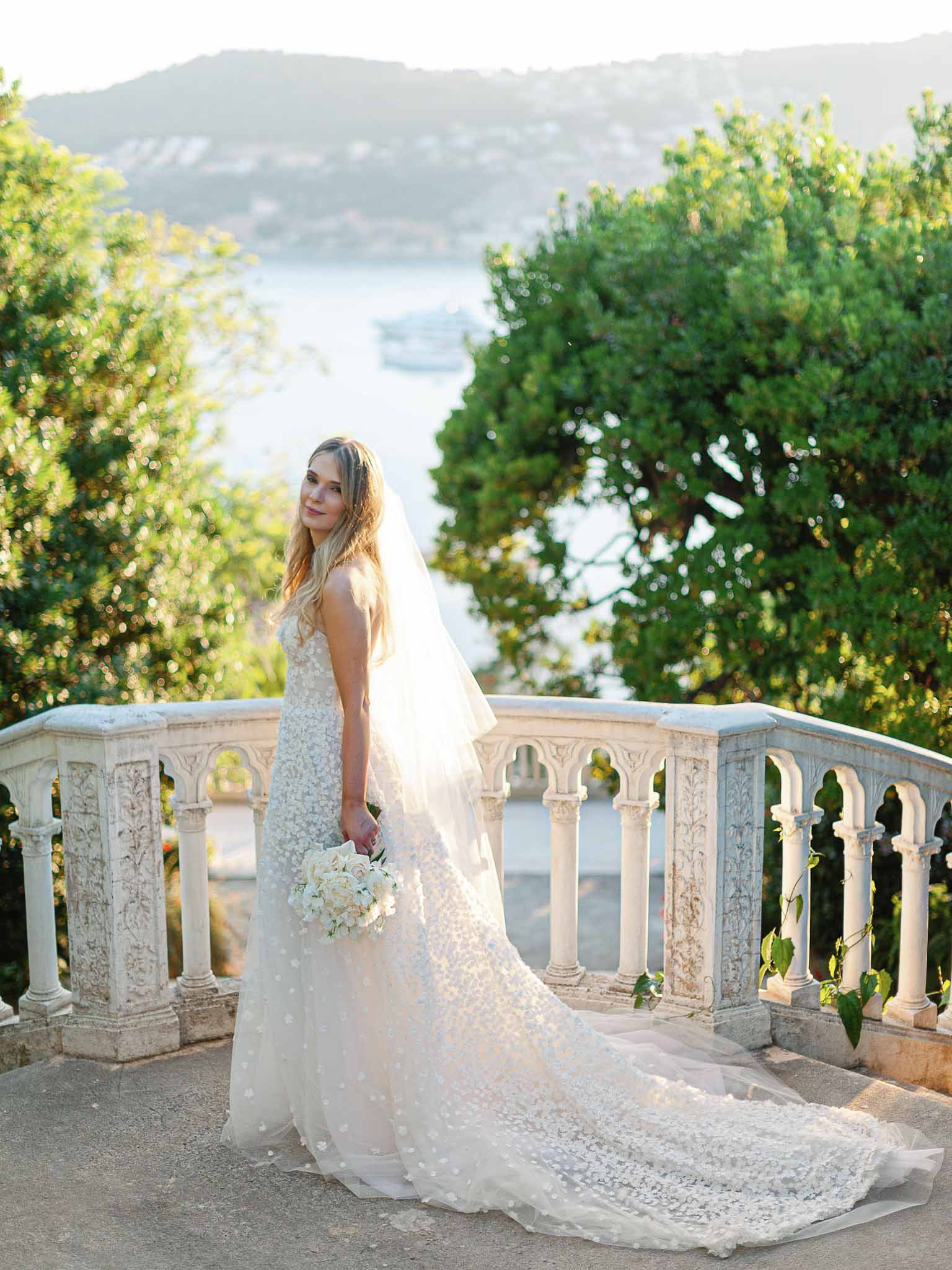 Bride in ivory lace gown on stone terrace balustrade overlooking lake at luxury wedding venue