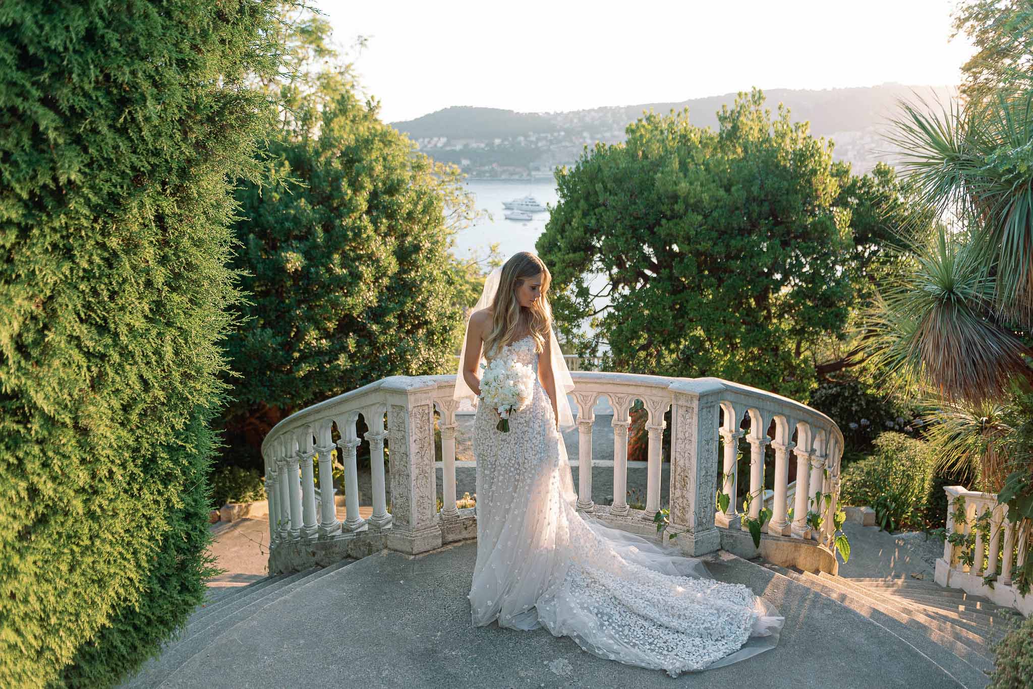 Bride in white lace gown standing on stone bridge at formal garden venue with lake view