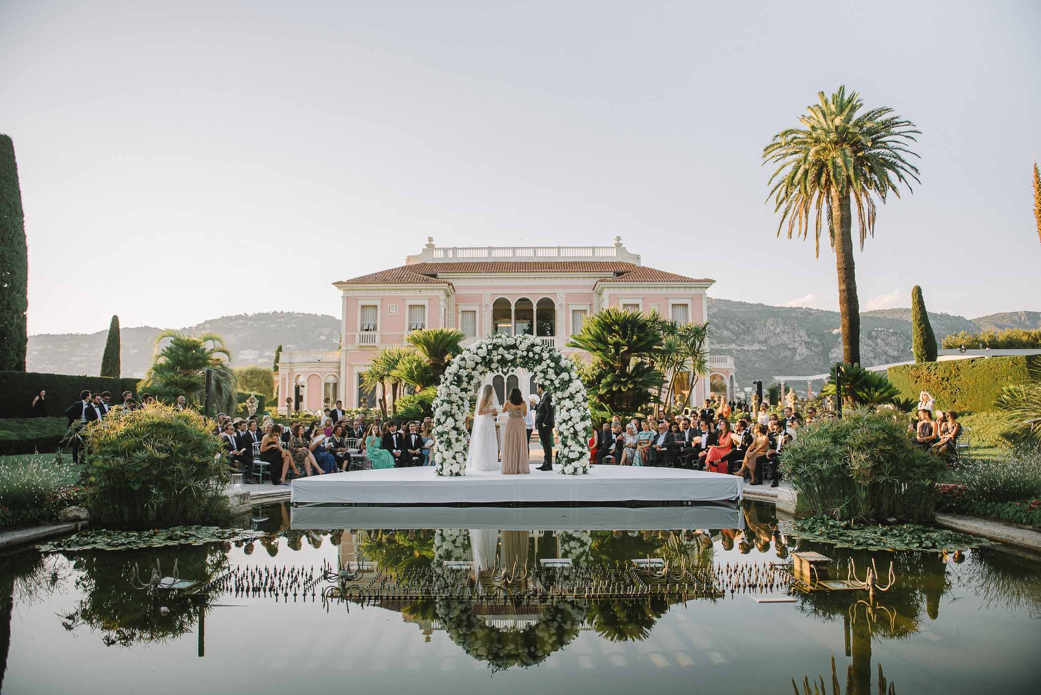Outdoor wedding ceremony on platform over pond at pink neoclassical villa with formal gardens