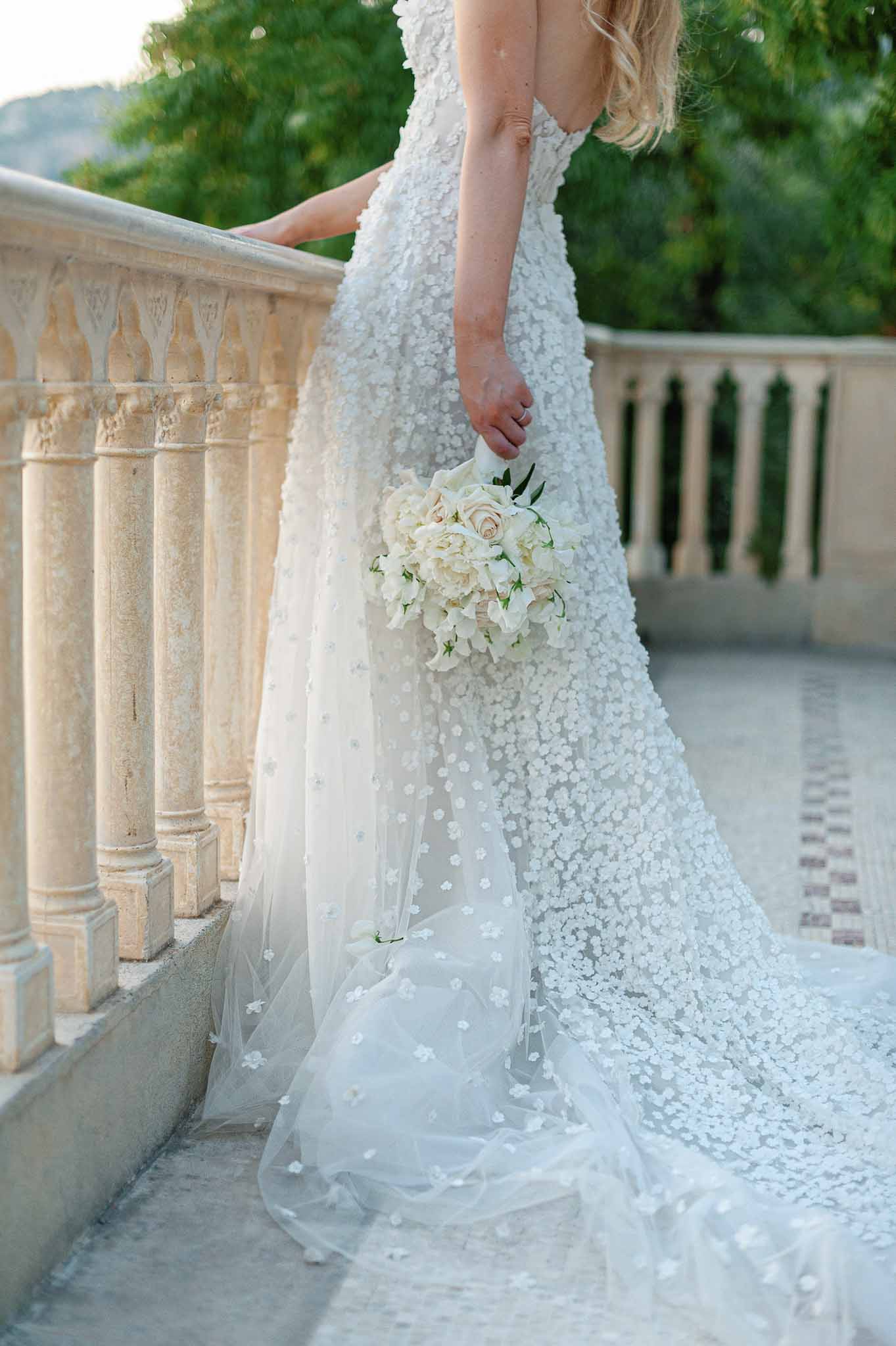 Bride in ivory beaded gown holding cream rose bouquet on stone terrace overlooking manicured gardens
